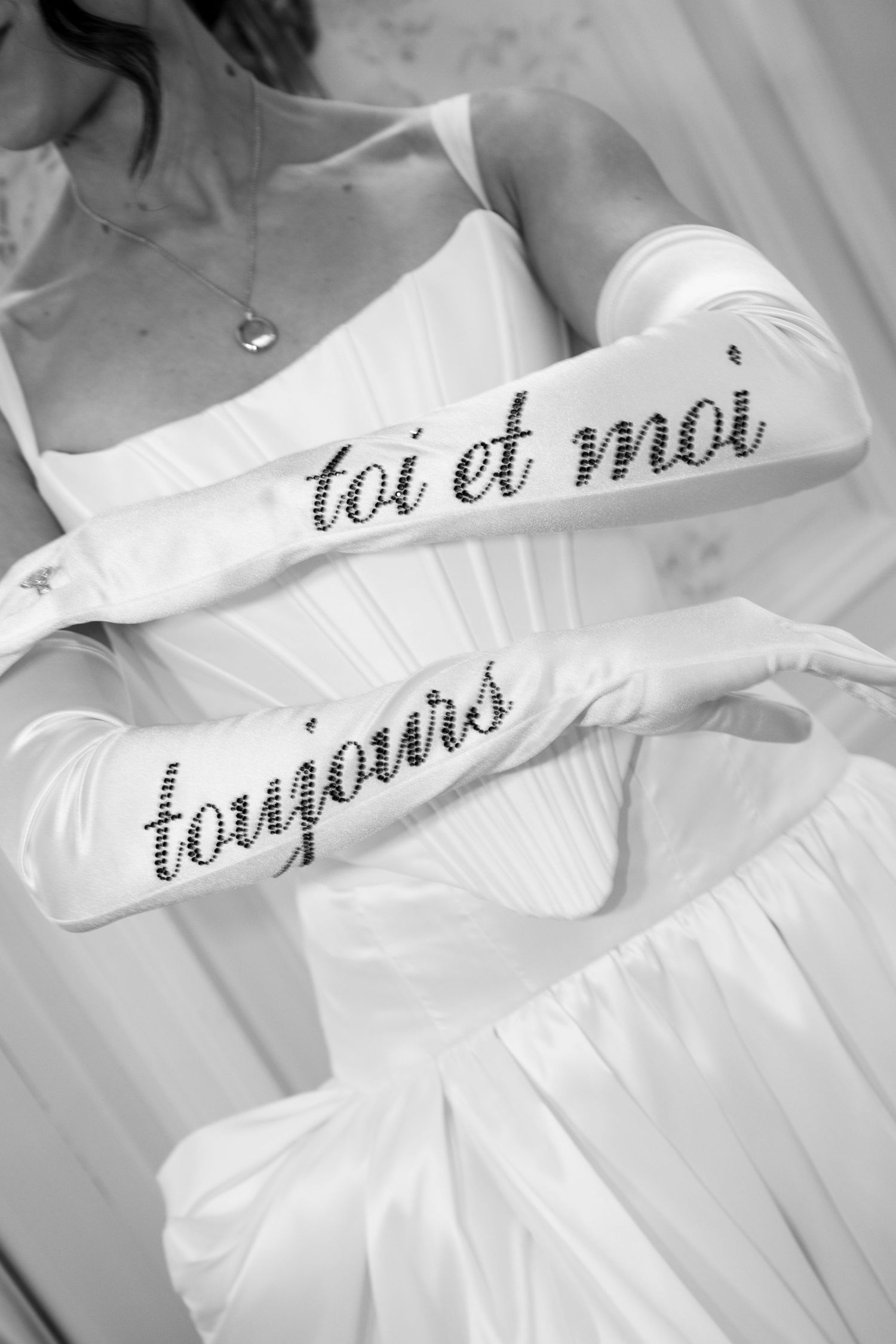 Black and white close-up of bride holding white satin gloves reading toi et moi toujours in rhinestones.