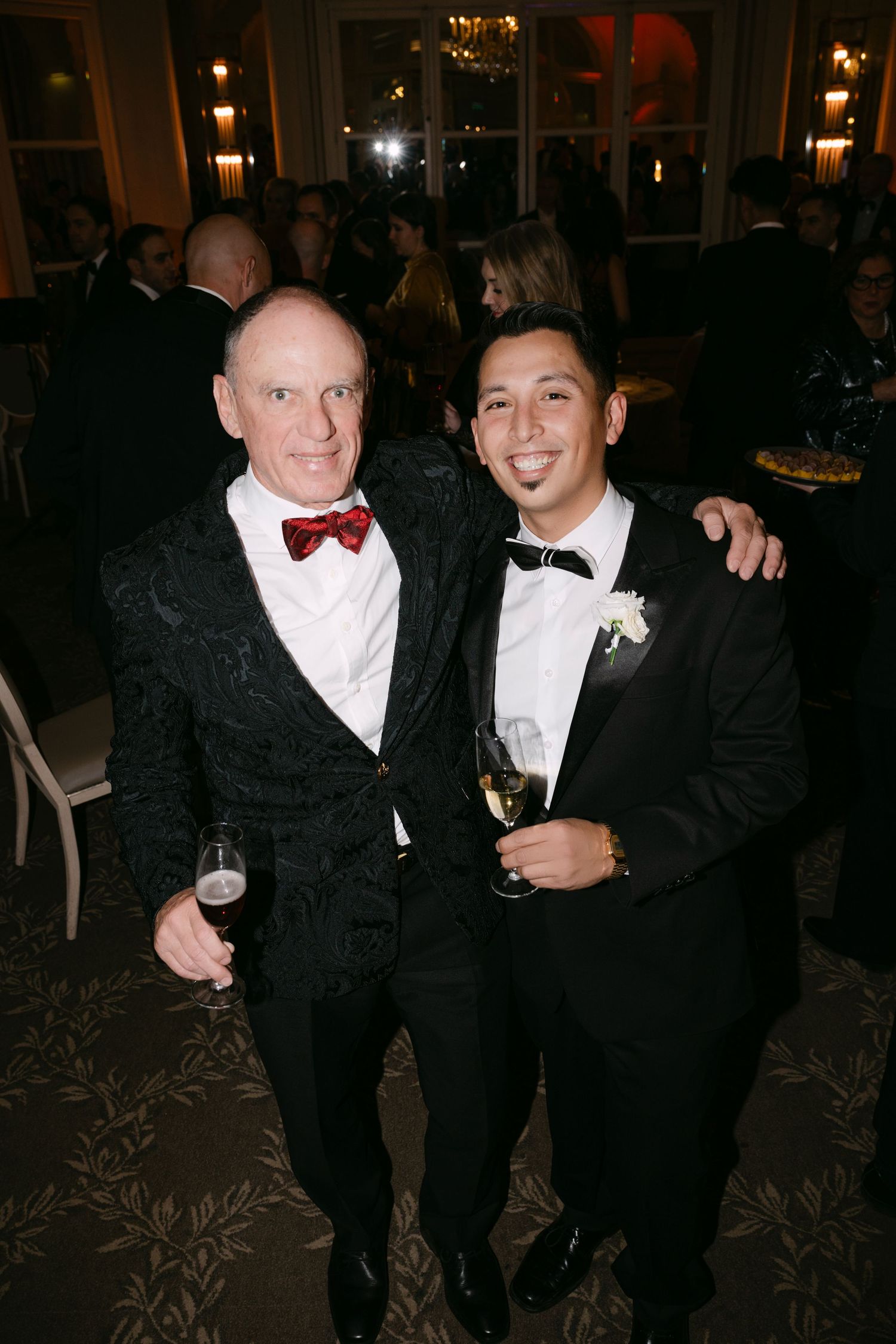 Two men in formal tuxedos smiling and holding champagne glasses at elegant evening wedding reception.