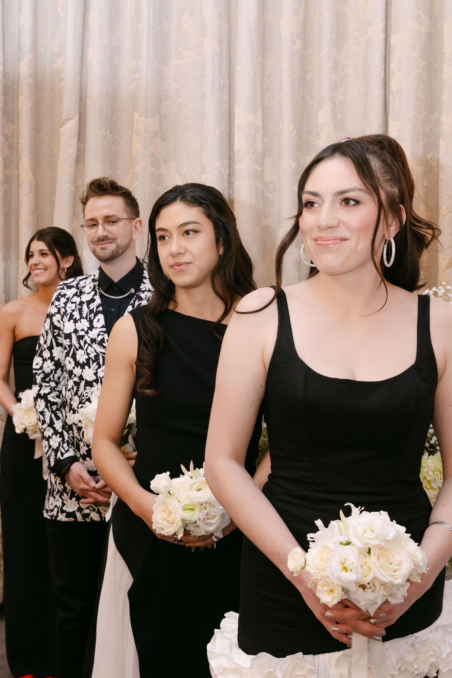 Bridesmaids in black dresses holding white bouquets walking down the aisle during an elegant wedding ceremony.