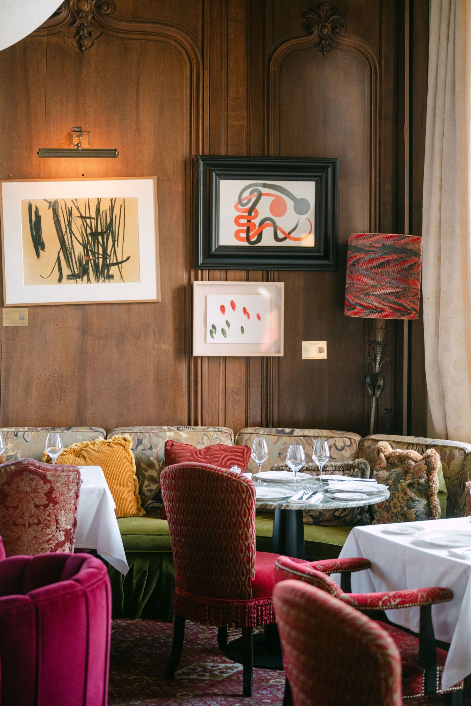 Sophisticated restaurant corner with wood-paneled walls, colorful velvet chairs, framed artwork, and intimate table setting.