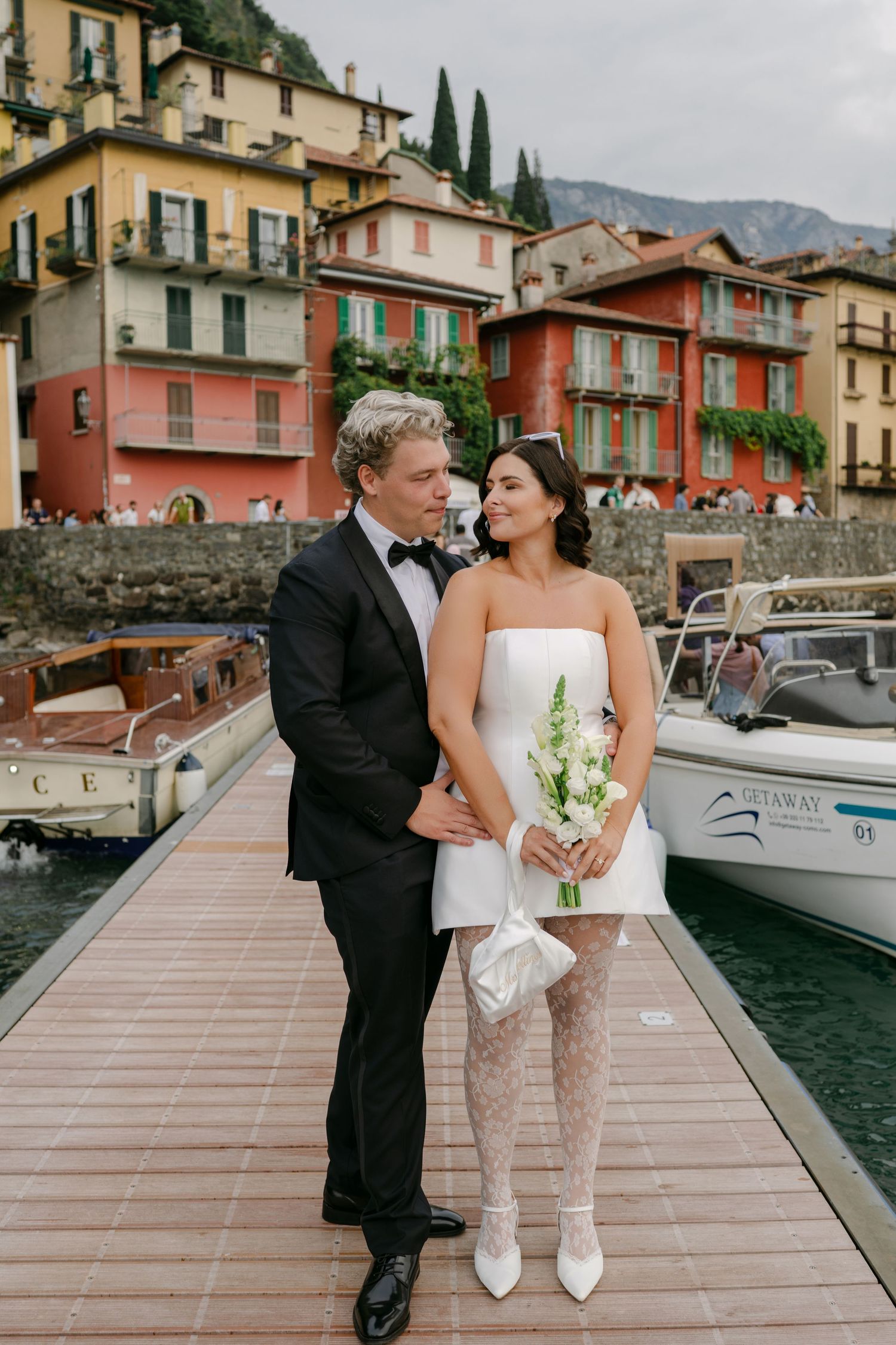 Bride and groom pose on a dock in Varenna, Lake Como, Italy, with colorful waterfront buildings behind them.