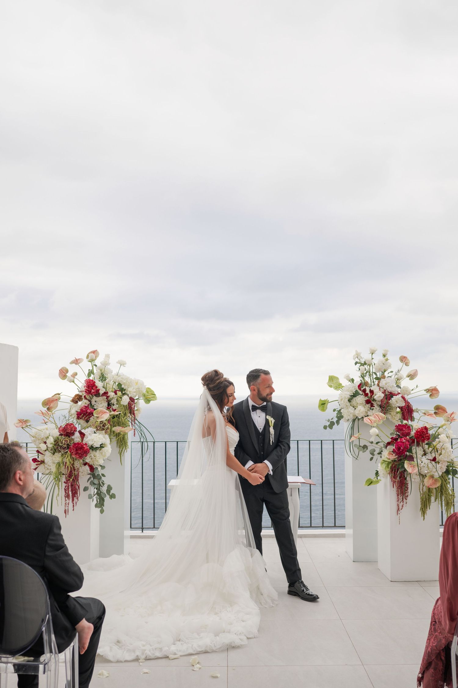 Bride and groom exchange vows at an outdoor wedding ceremony with floral arrangements overlooking the ocean.