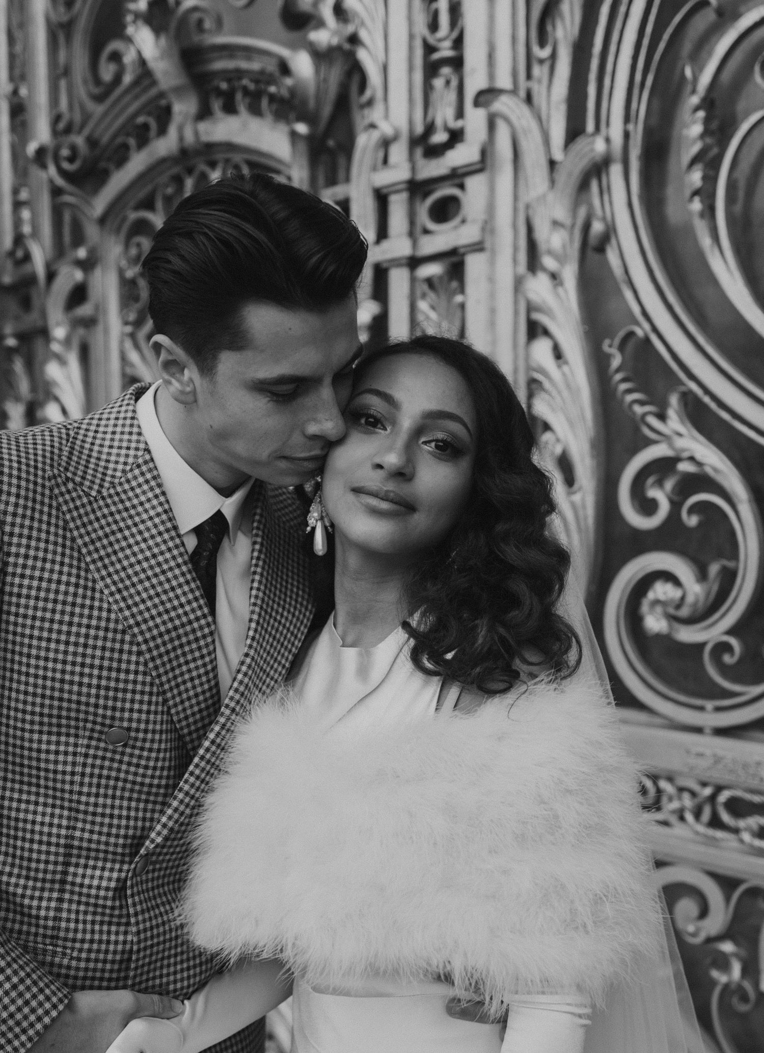 A black and white portrait of newlyweds posing against decorative architecture.