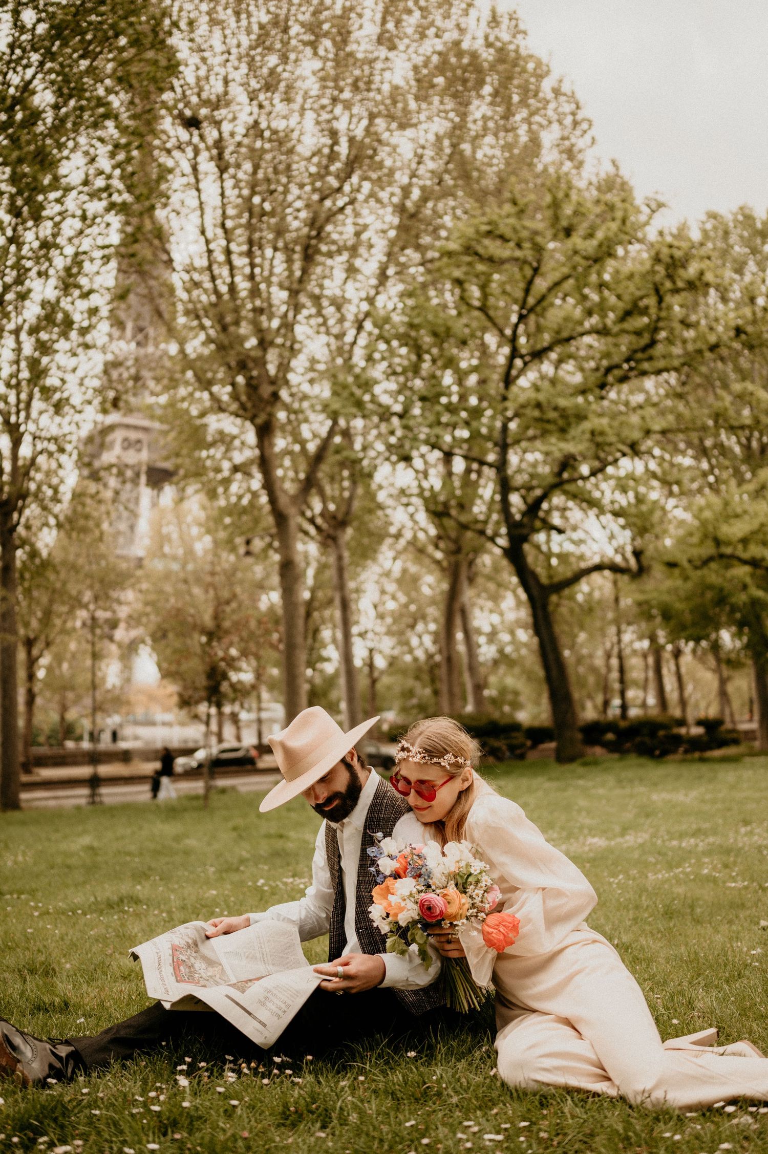 Couple in vintage clothing reading together on park lawn with spring trees and Eiffel Tower behind.