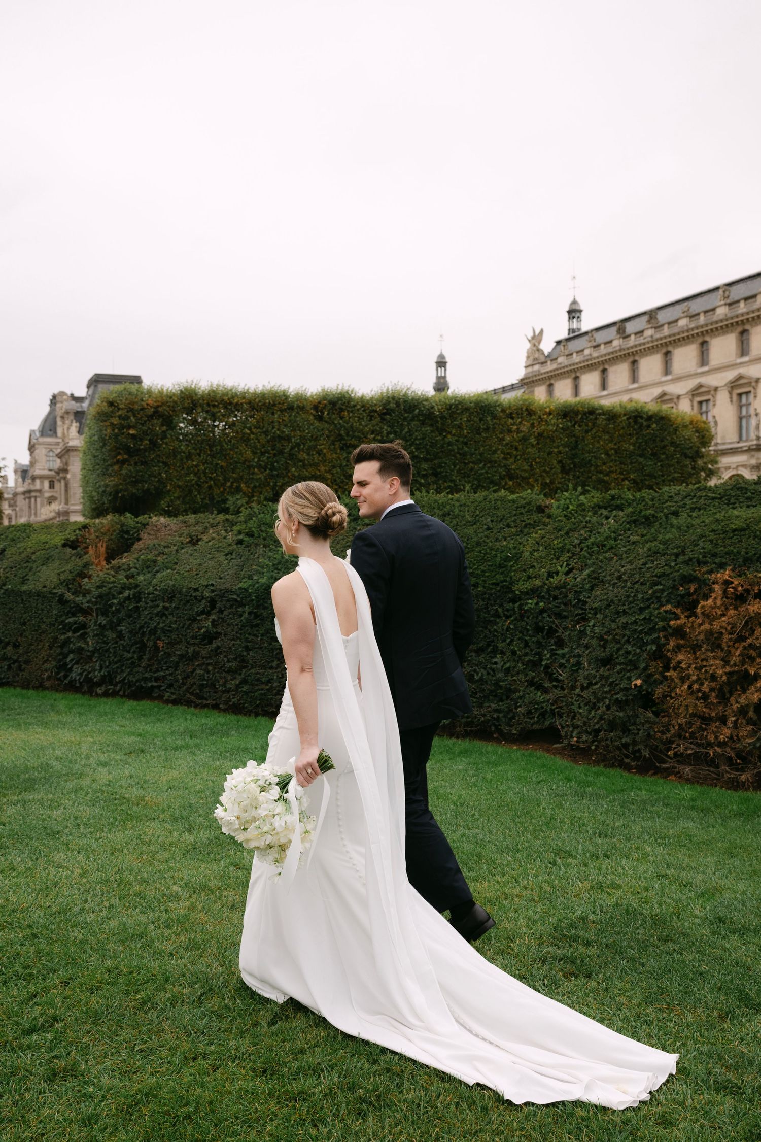 Wedding couple standing in formal garden with manicured hedges and historic building backdrop.