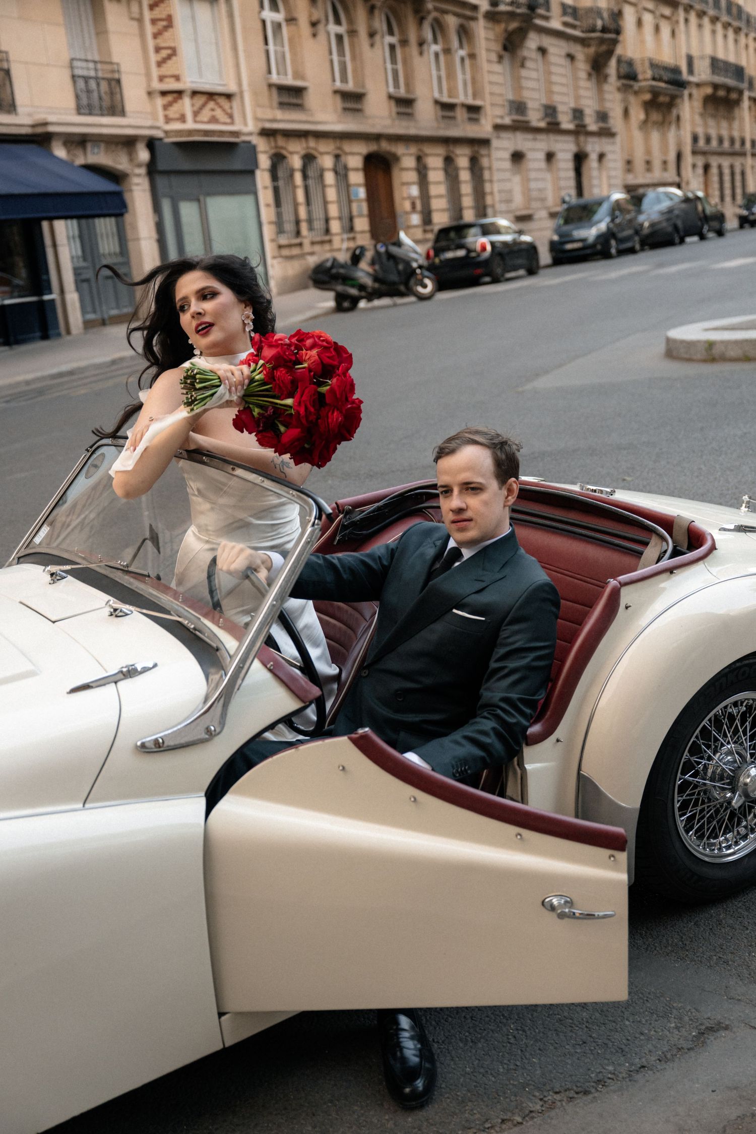 A couple enjoys a playful moment in a classic white convertible car on a Parisian street.