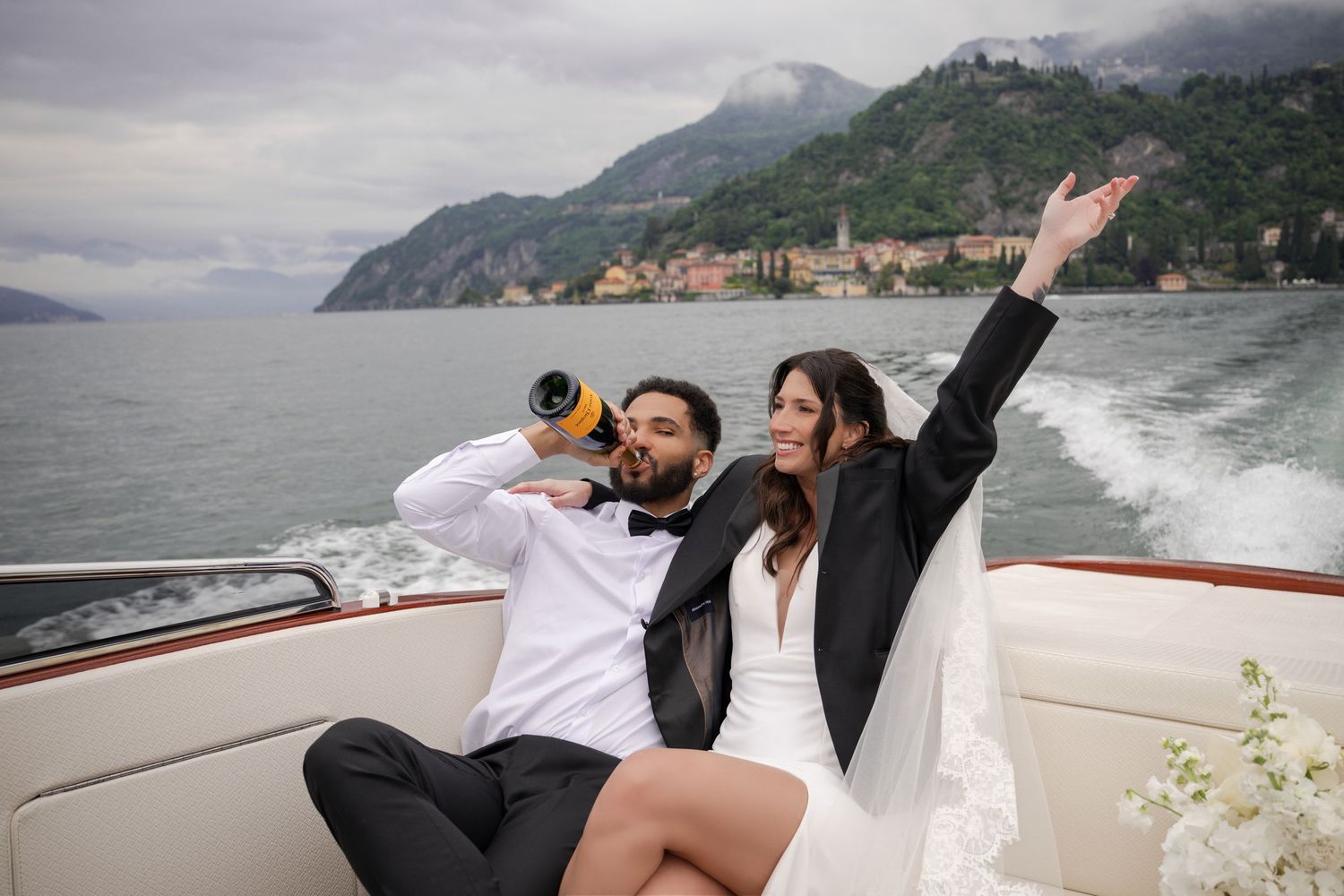 Couple celebrating on a boat on Lake Como, groom drinking champagne as bride raises her arm joyfully.