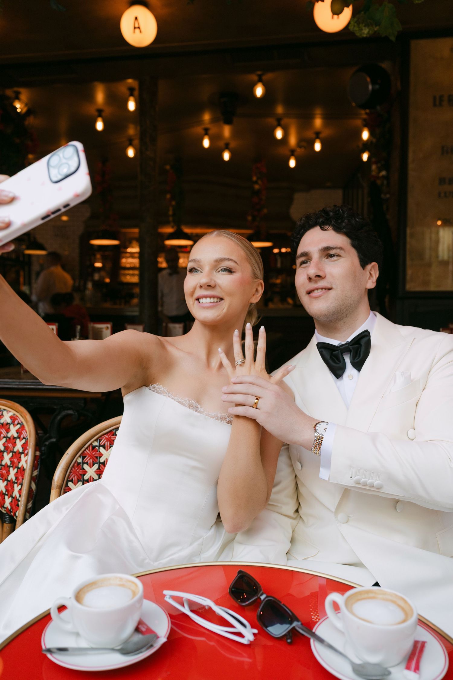 Newlyweds take a selfie at a Parisian cafe, bride showing off wedding ring while wearing white gown, groom in white tuxedo.