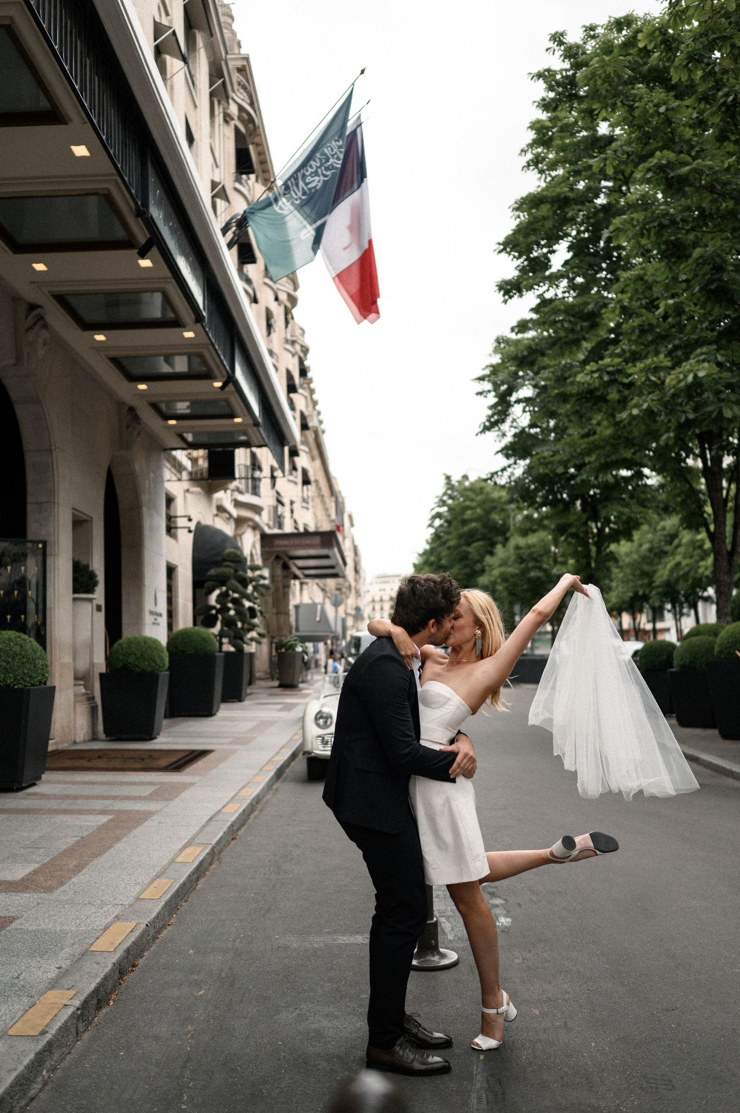 Couple sharing romantic kiss outside Four Seasons Paris, bride in mini dress lifting leg with veil flowing.