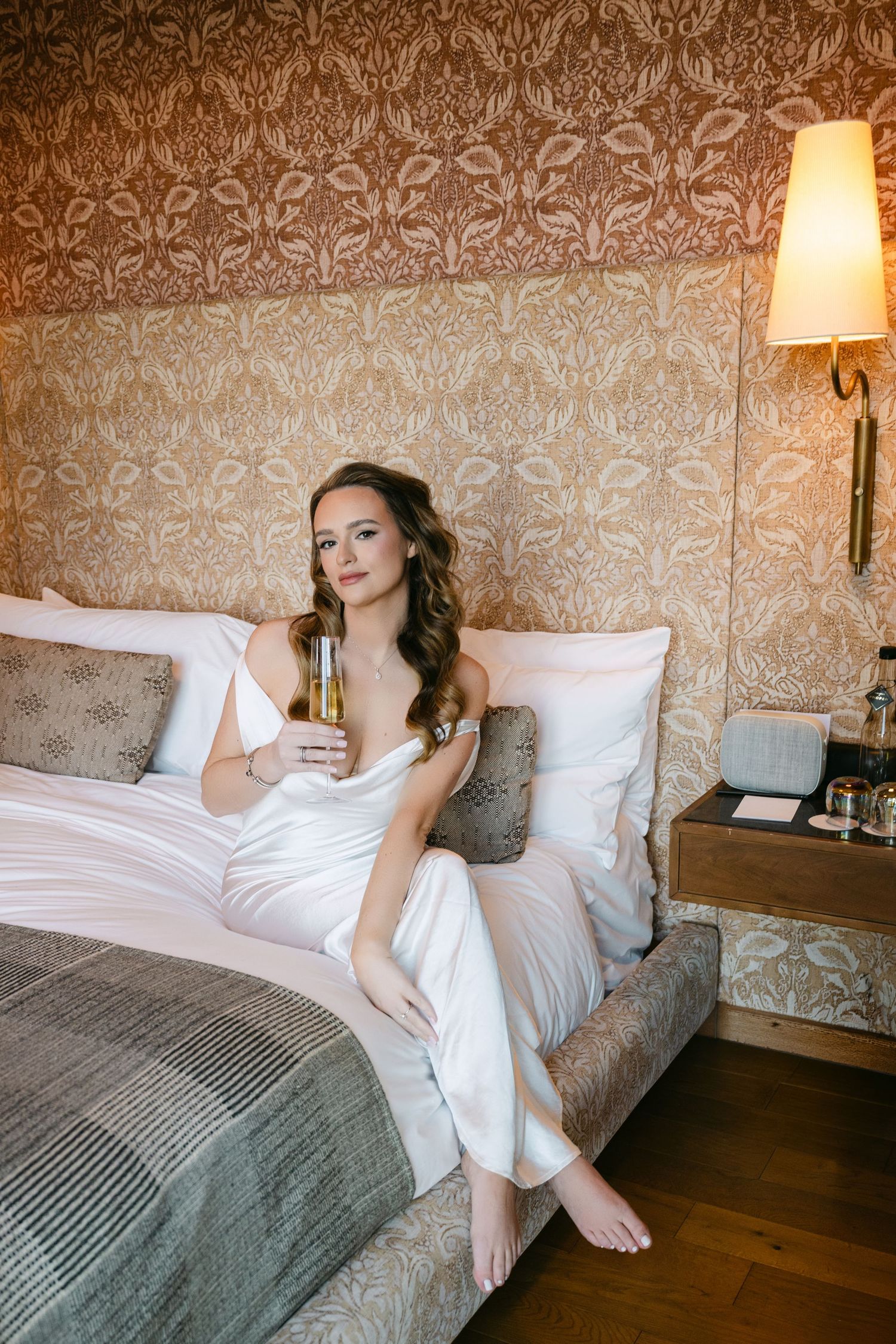 Beautiful bride in white satin dress sitting on hotel bed holding champagne glass with ornate wallpaper backdrop.