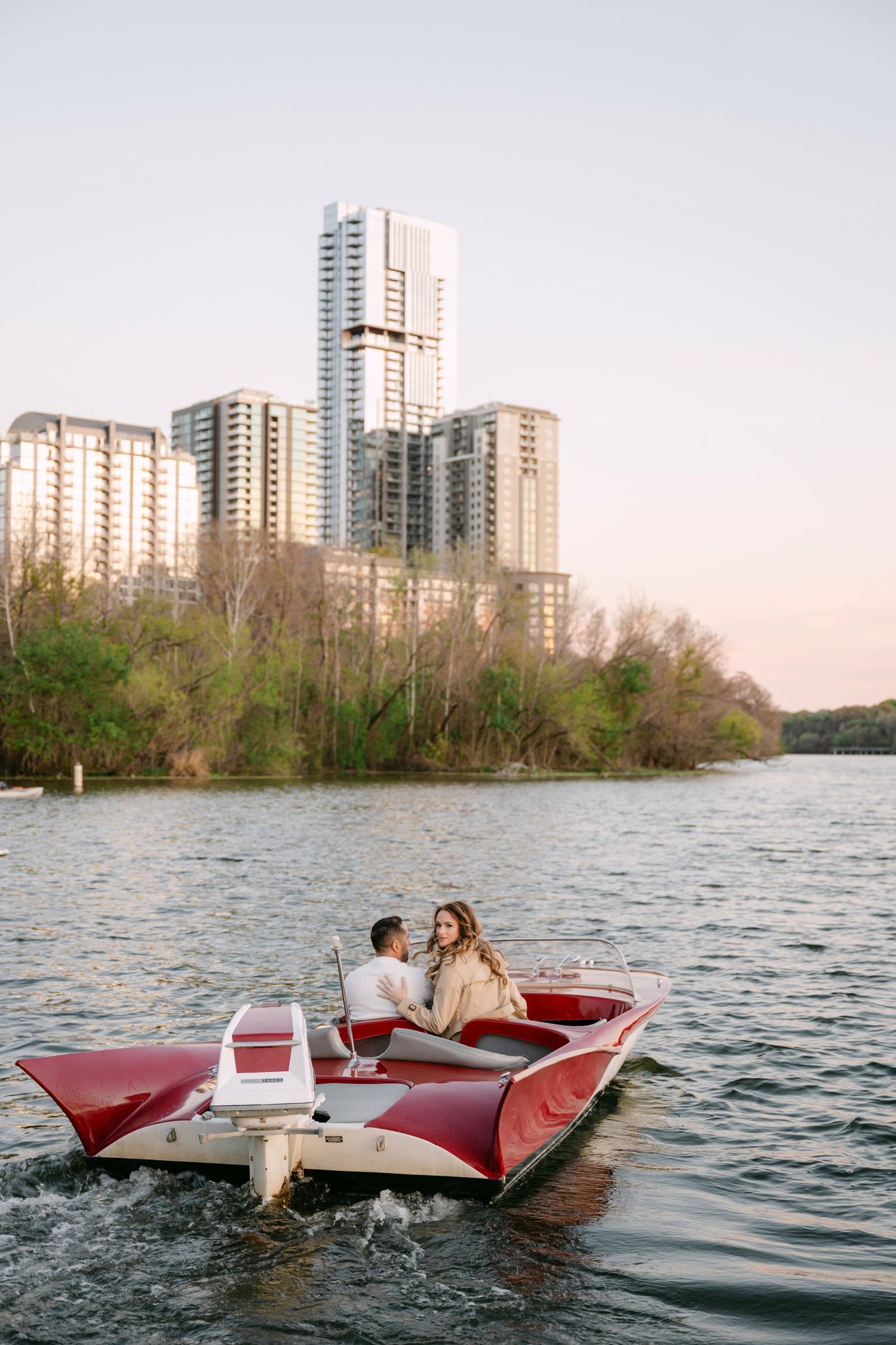 Couple riding a red speedboat on a river with city skyscrapers in background.