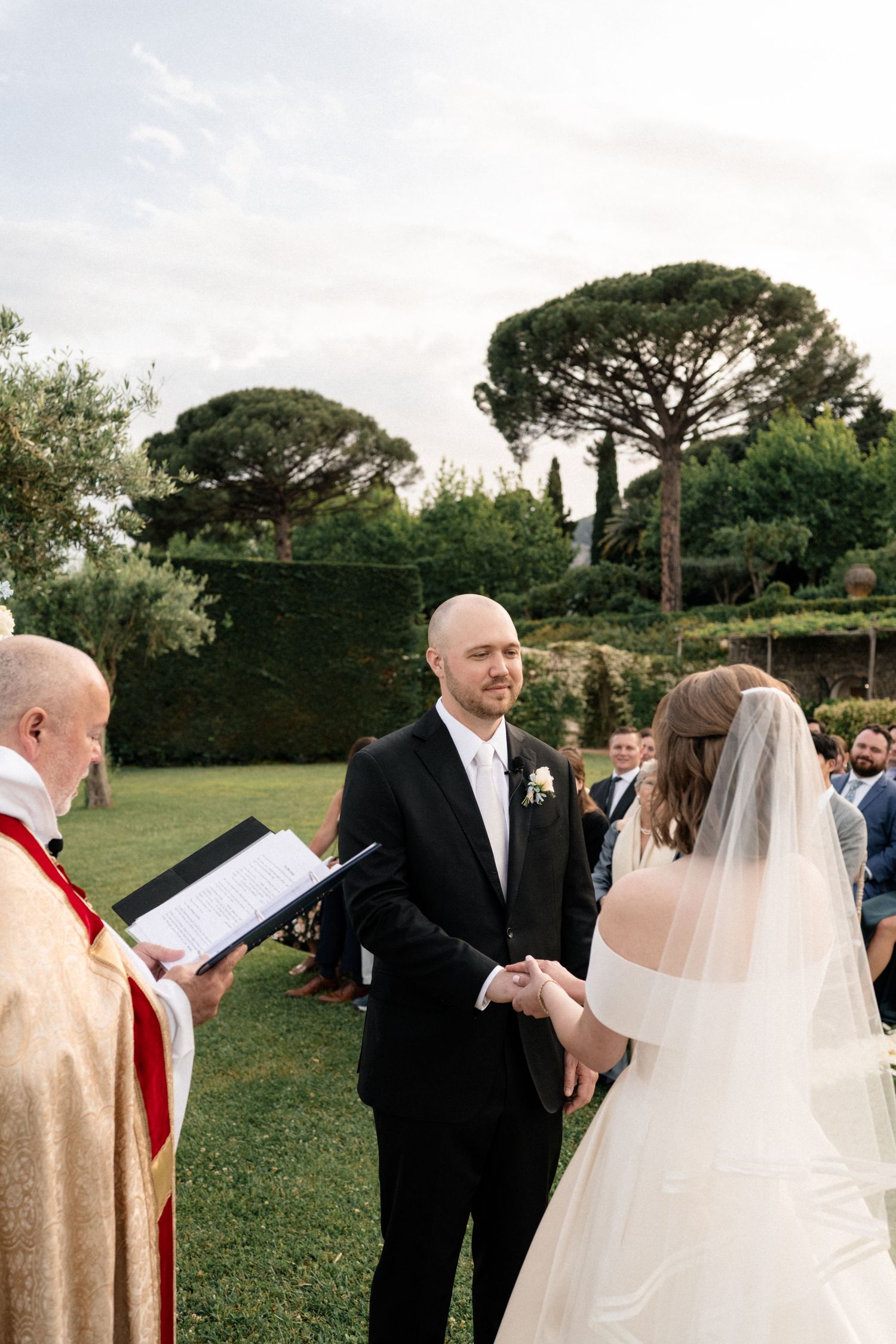 Groom smiles at bride during outdoor wedding vows with officiant in red vestments.