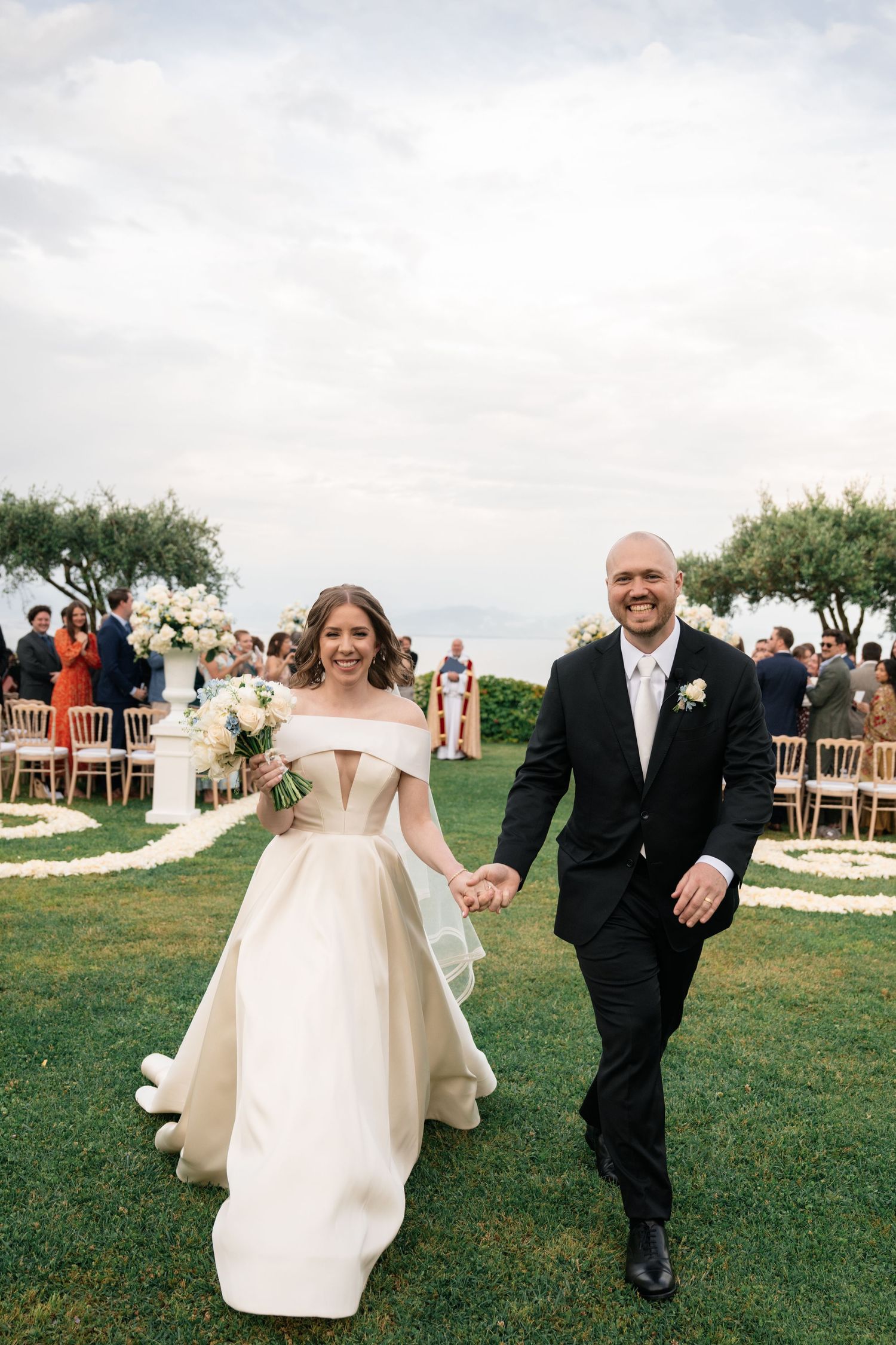 Smiling newlyweds walk down aisle hand in hand after outdoor wedding ceremony with white floral arrangements.