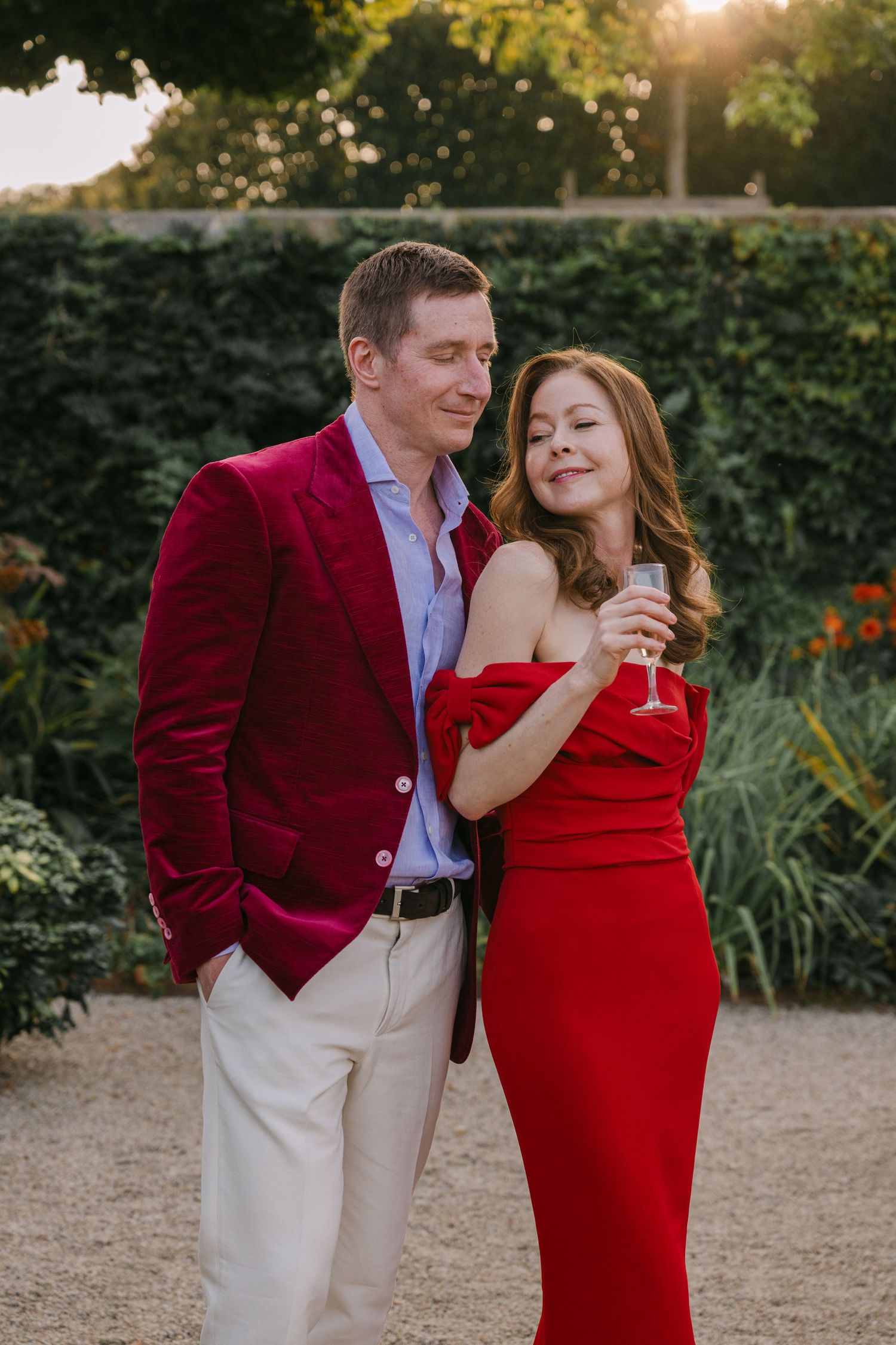 Couple embracing at golden hour in garden, man in burgundy velvet blazer, woman in red dress.