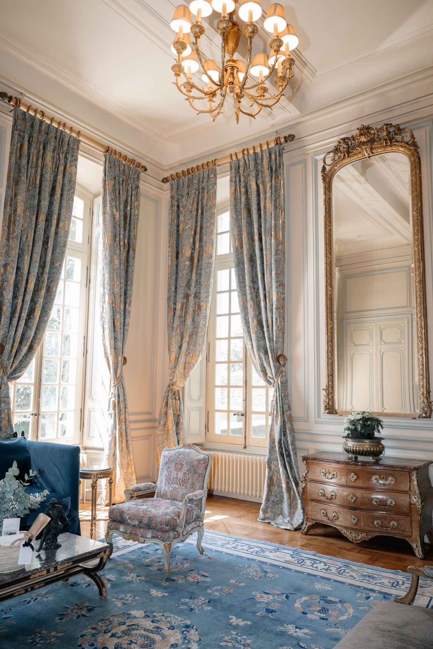 Elegant French chateau room with blue damask curtains, gilded mirror, antique armchair, and chandelier.