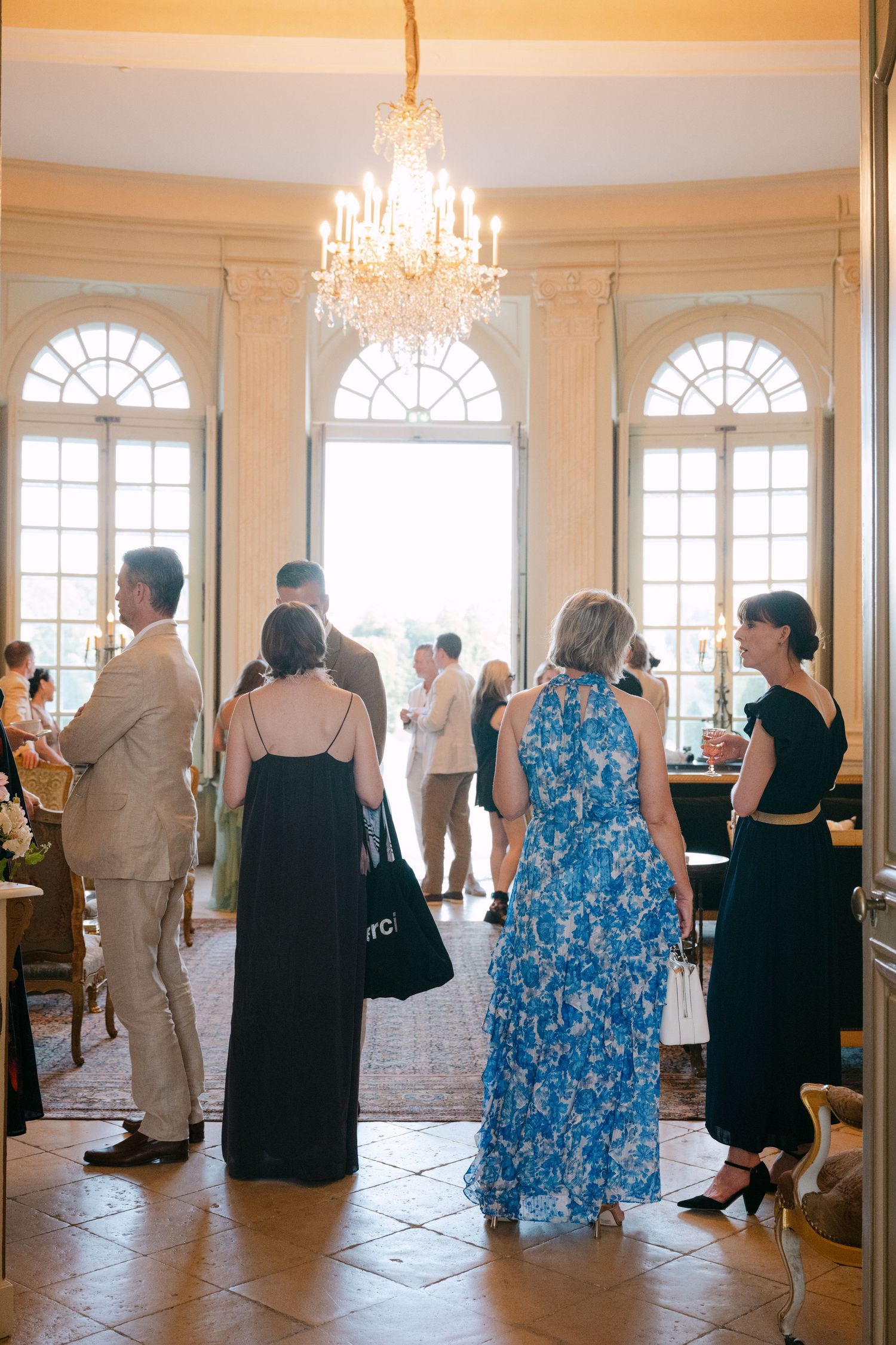 Elegantly dressed wedding guests gather near arched windows under a crystal chandelier in a grand venue.