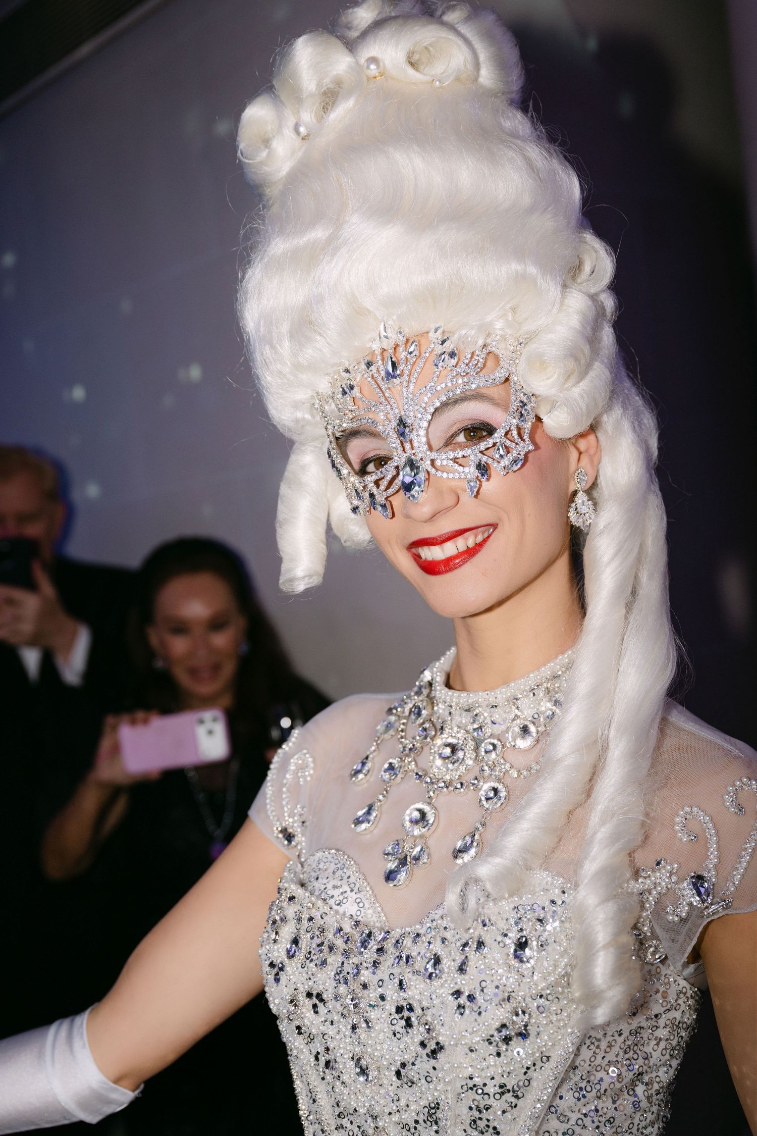 Elegant performer in sparkling silver costume with ornate jeweled mask and towering white wig at a gala event.