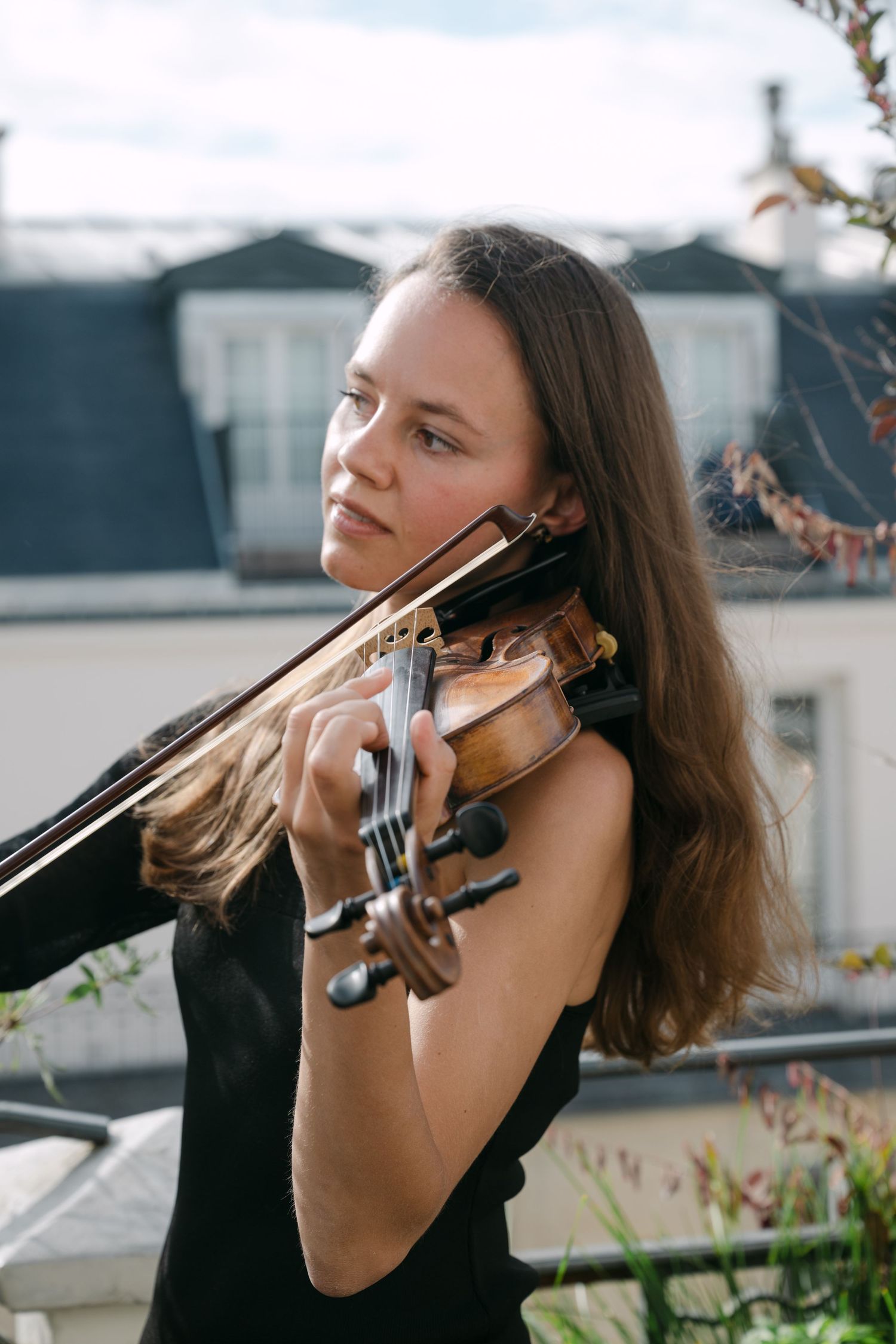 Female violinist in black dress performs outdoors on Parisian rooftop at wedding ceremony.