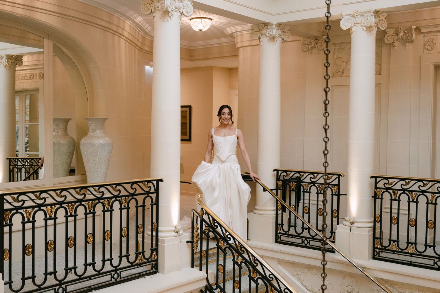 Bride in sleek white gown descending grand staircase with black iron railings in luxury venue.