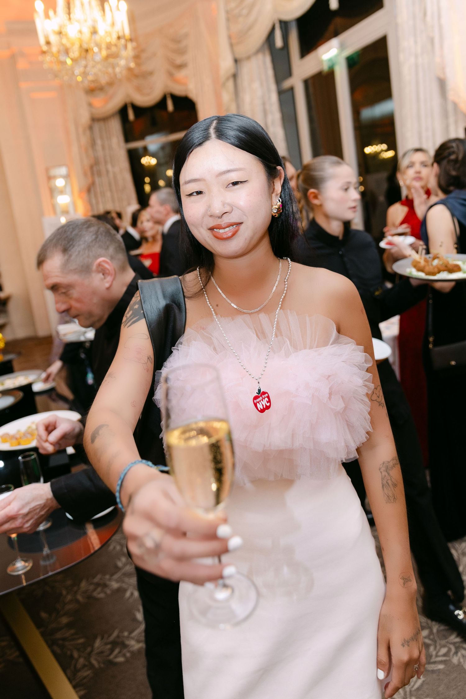 Asian woman in pink ruffled off-shoulder dress holding champagne flute at upscale party venue.
