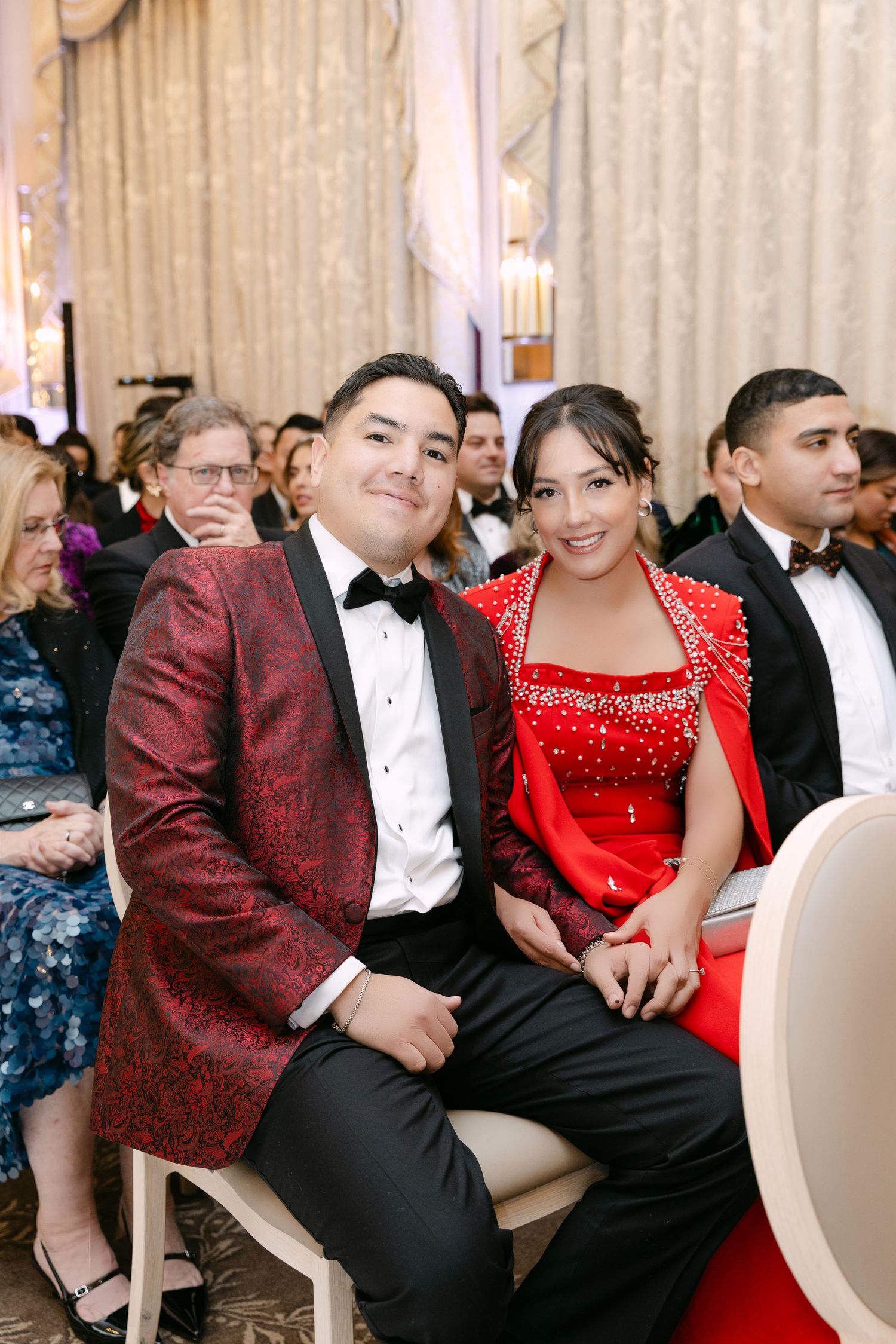 Couple in matching red and burgundy formal attire smiling together at a wedding ceremony in a grand ballroom.