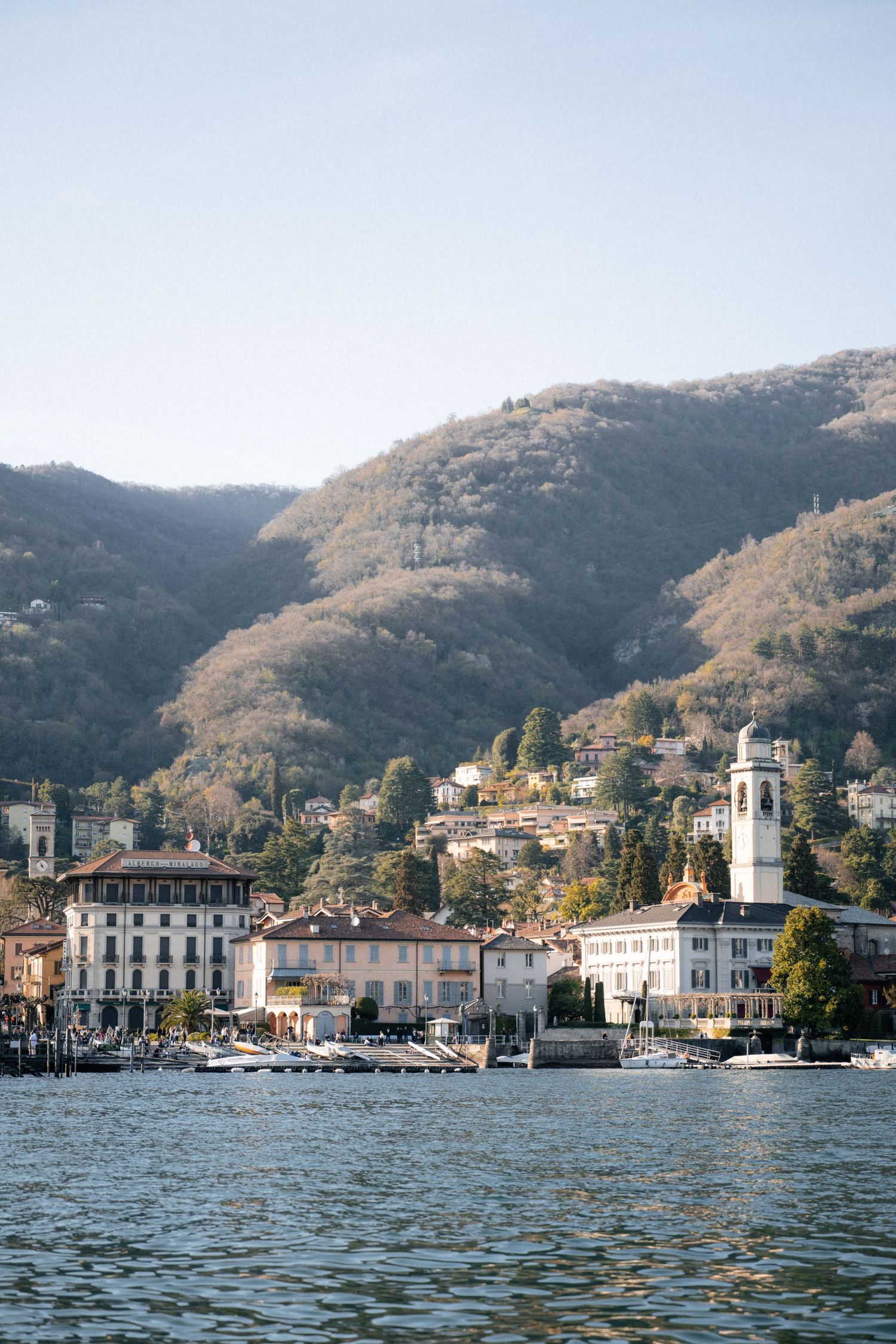 Picturesque Lake Como waterfront village with bell tower and lush green mountains under clear sky.