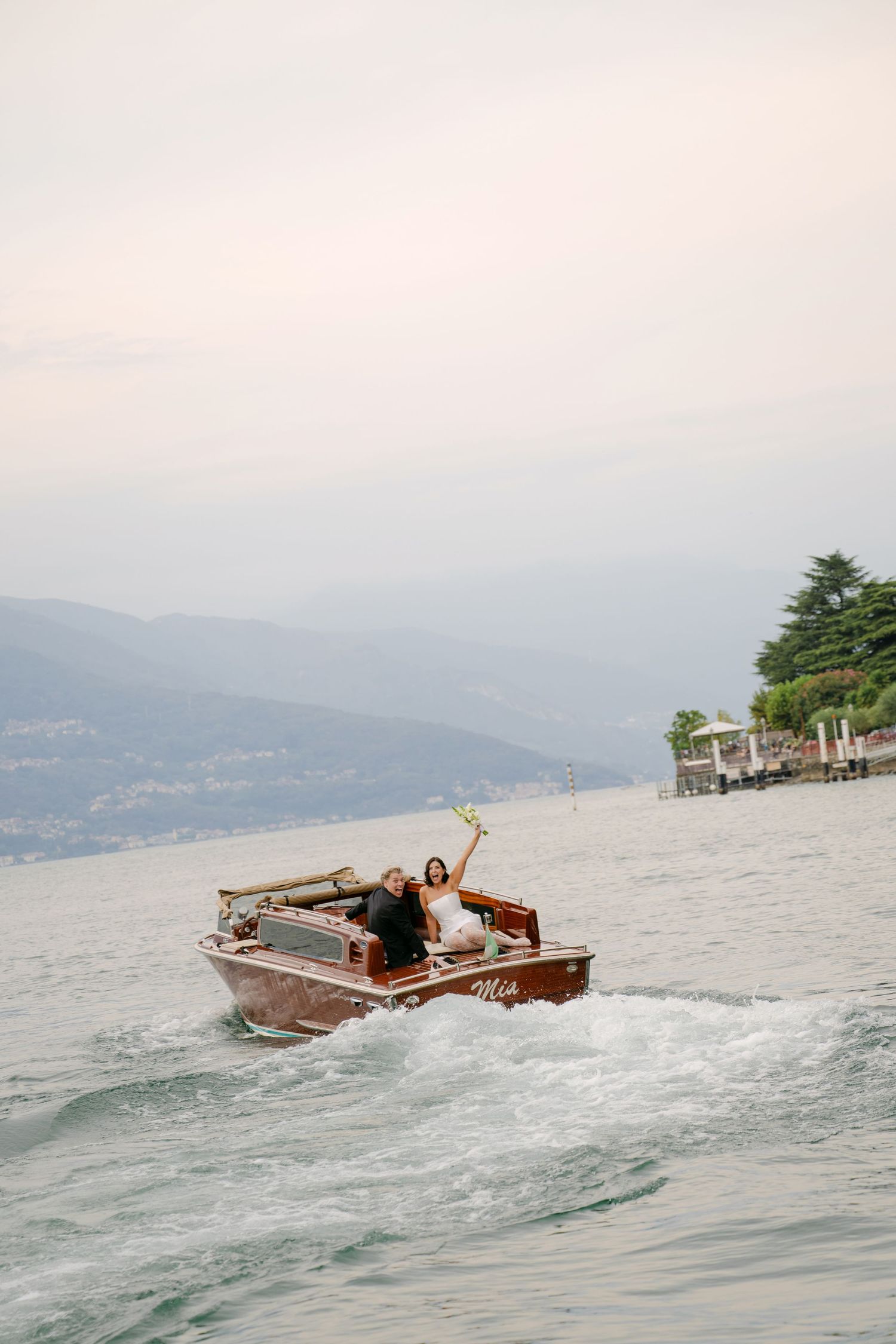 Bride and groom celebrating on a classic wooden boat speeding across a scenic Italian lake on their wedding day.