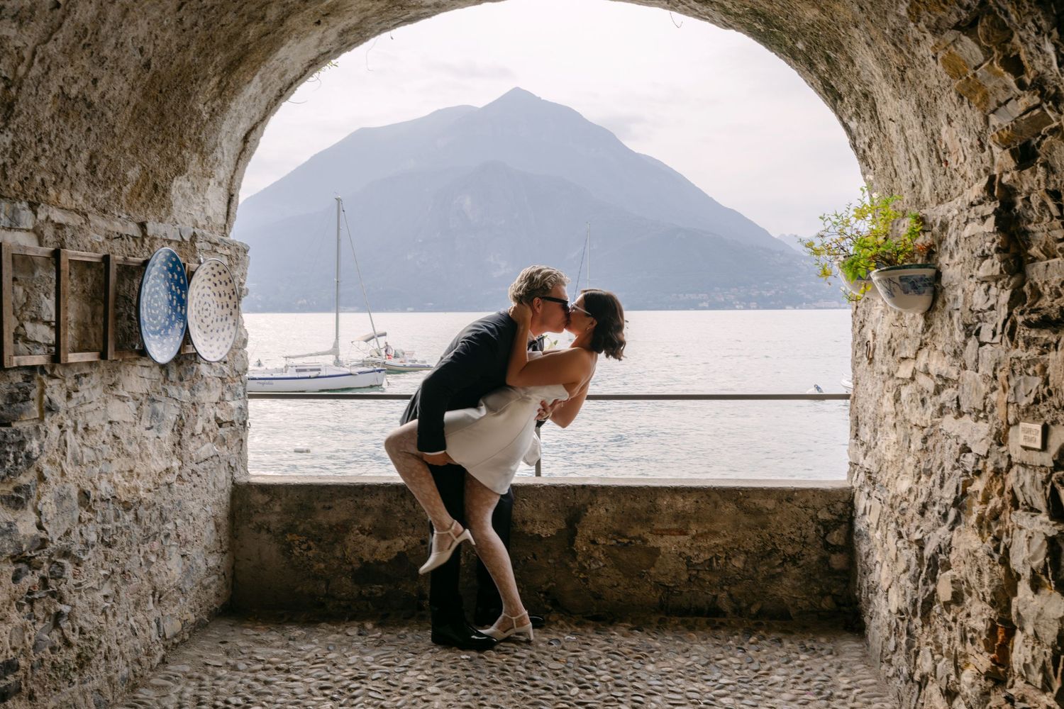 Couple sharing a romantic dip kiss under a stone arch overlooking Lake Como with mountain views.