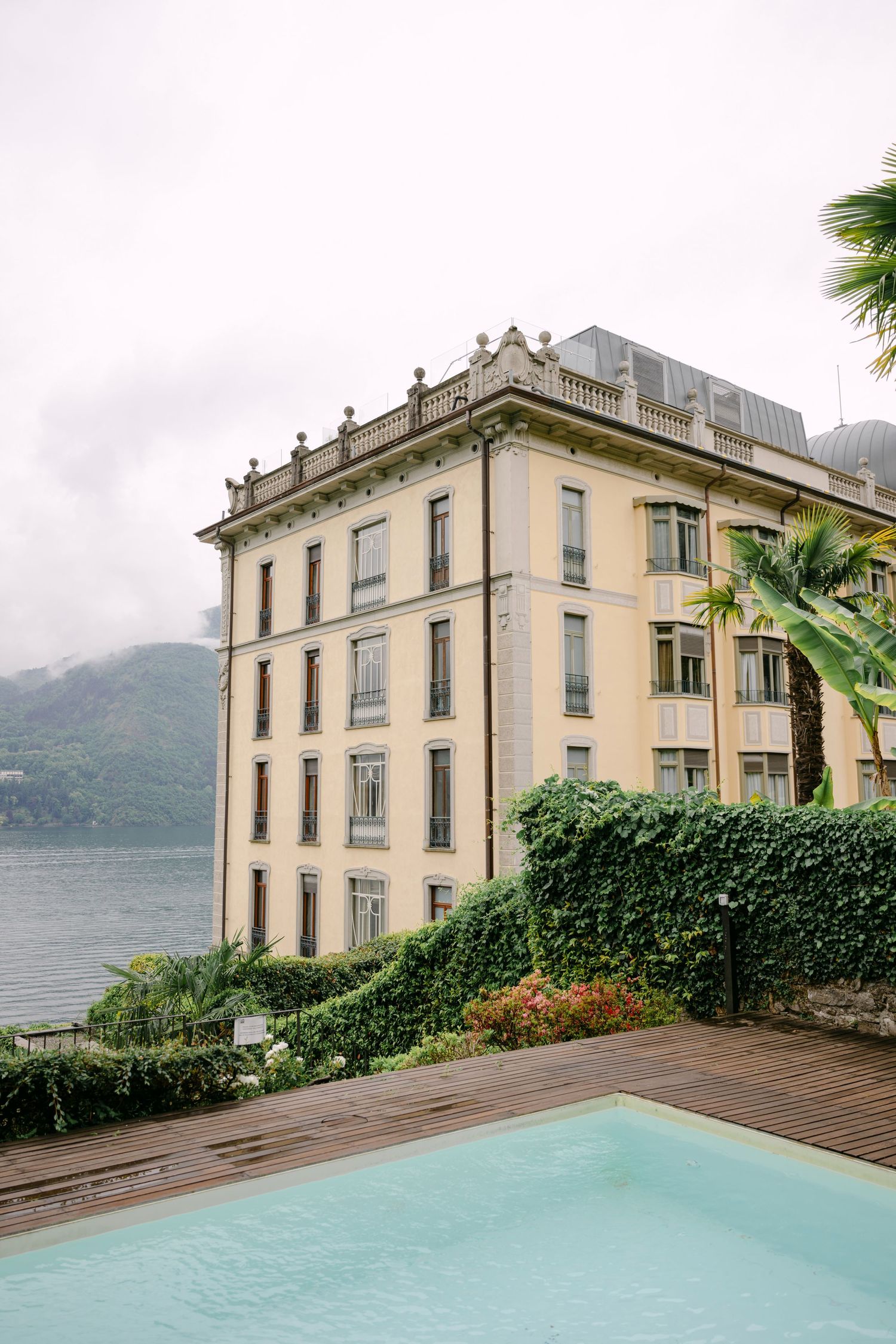 Elegant Italian villa with pool overlooking misty Lake Como, surrounded by lush greenery and palm trees.