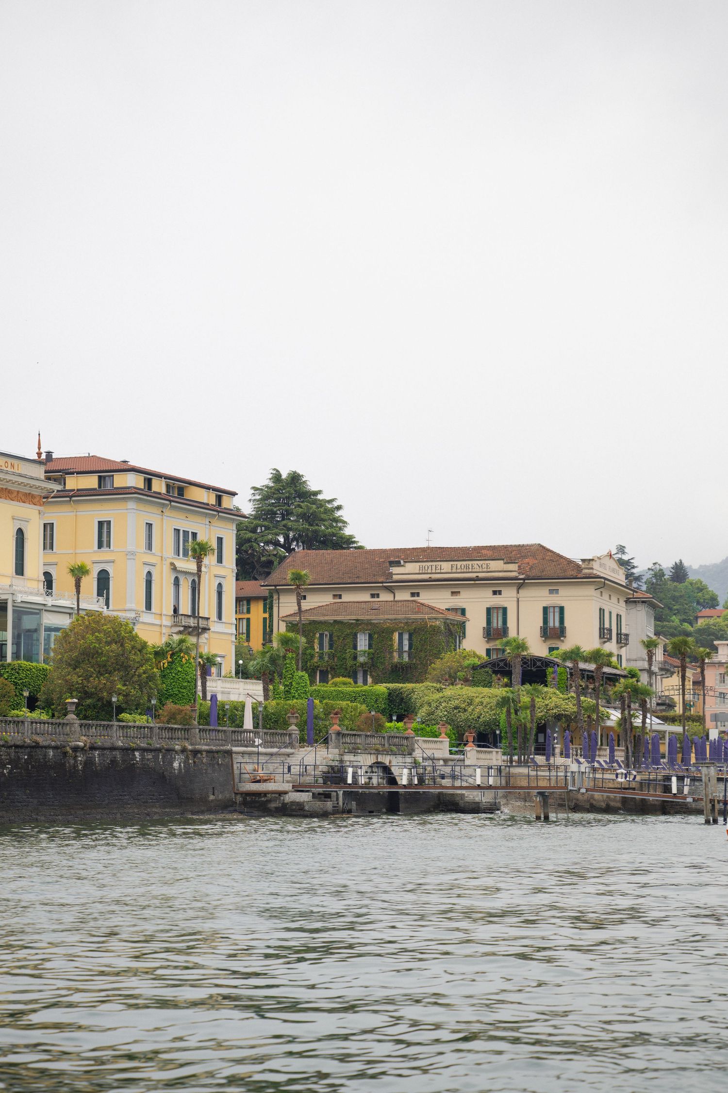 Elegant Italian lakeside villas with lush gardens reflected along calm misty Lake Como waterfront.