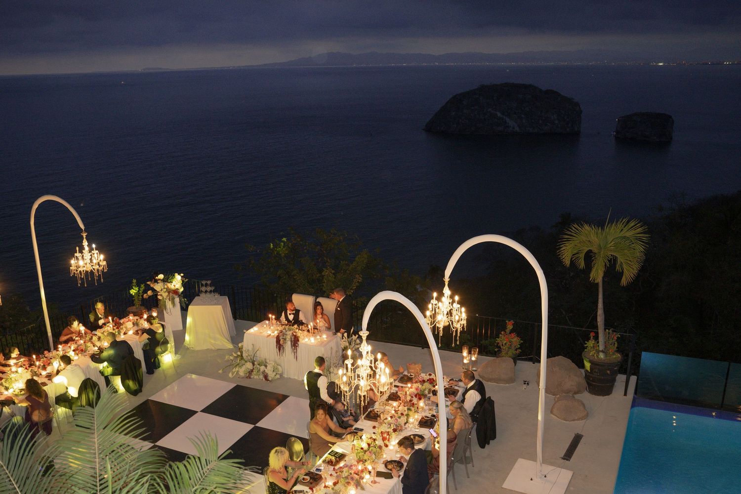 Elegant outdoor cliffside dinner party with chandeliers and pool overlooking ocean at night.