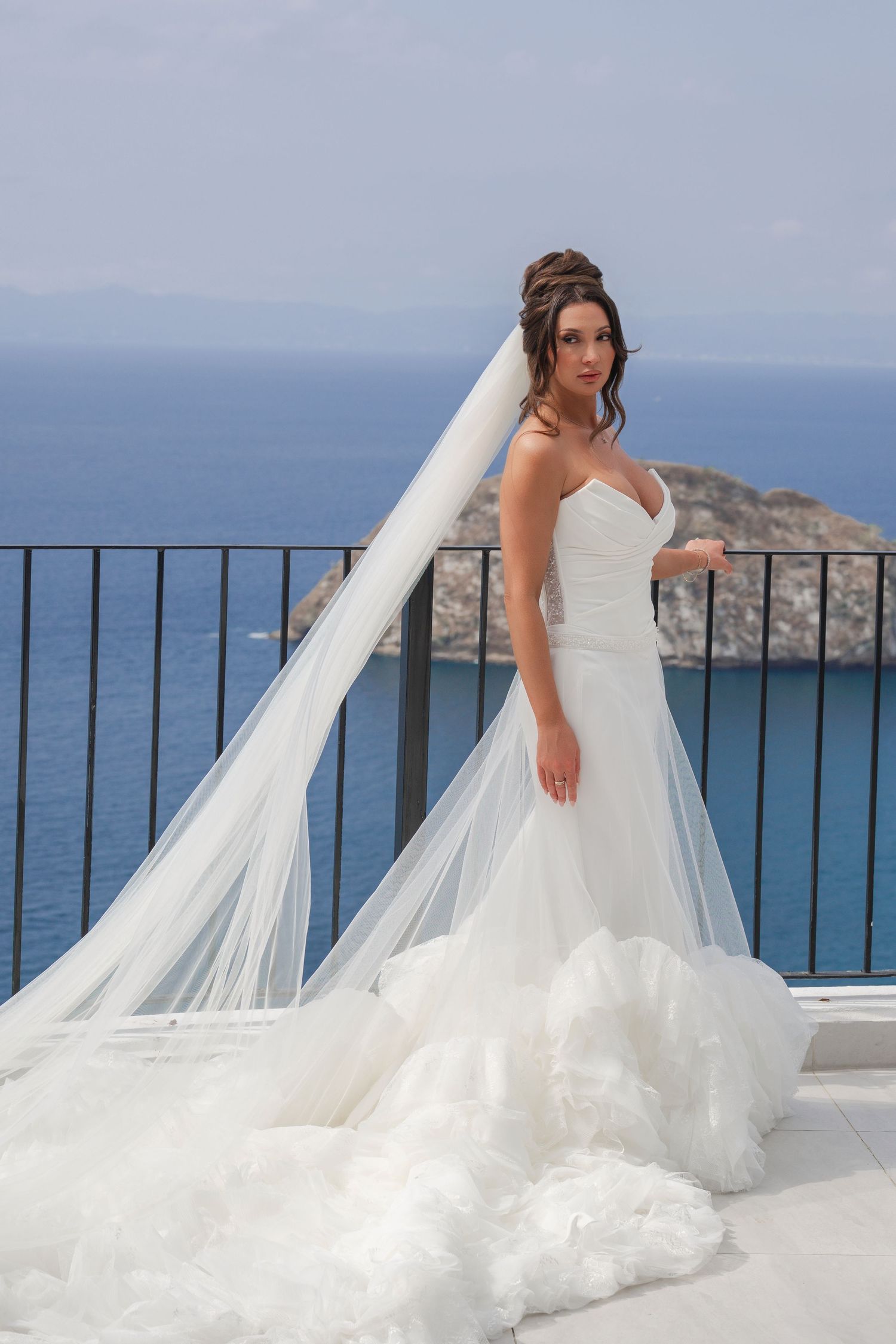 Bride in white strapless mermaid gown with long veil stands on seaside balcony with blue ocean and island backdrop.