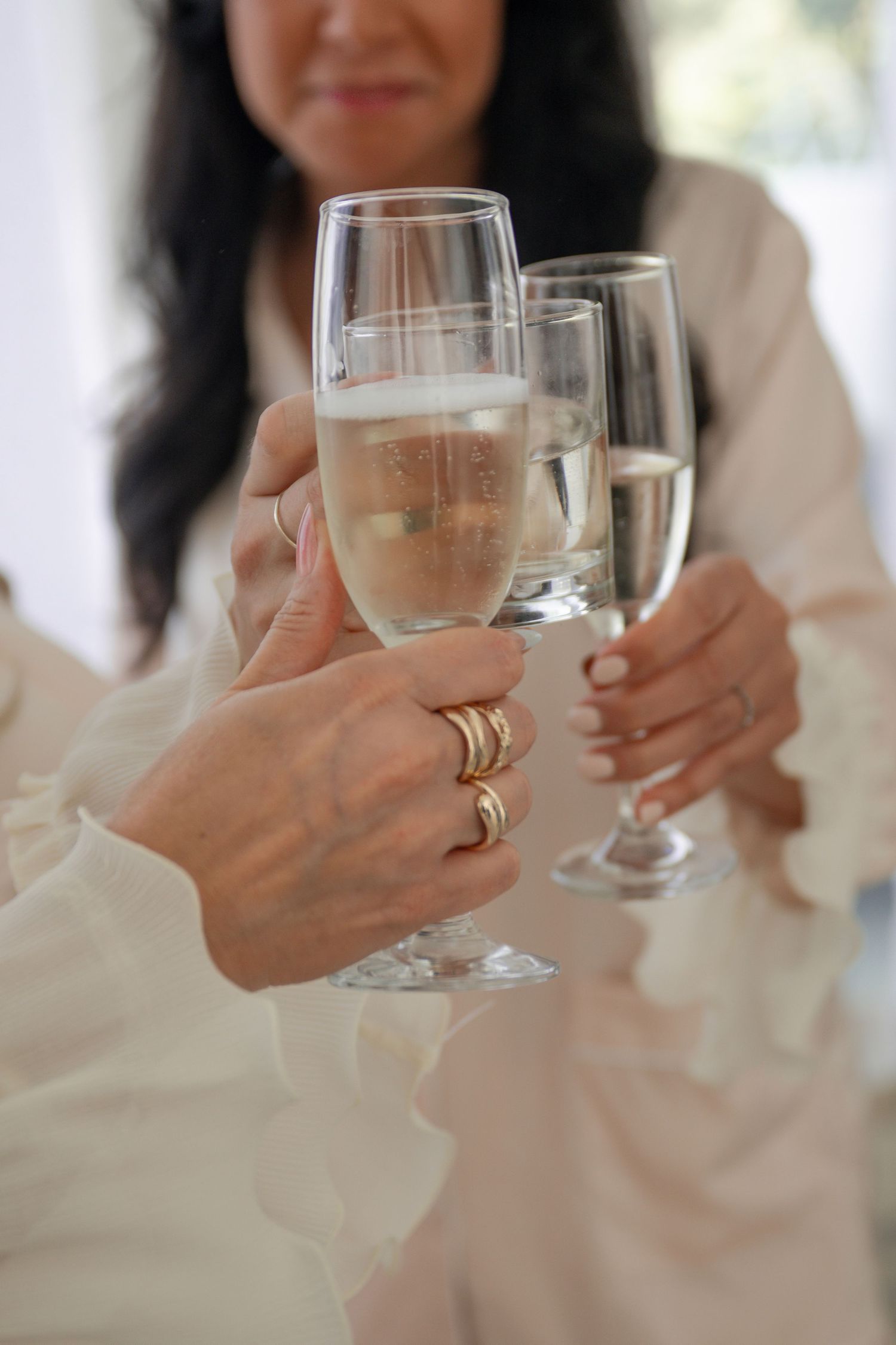 Women toasting with champagne flutes, hands adorned with gold rings, wearing white robes.
