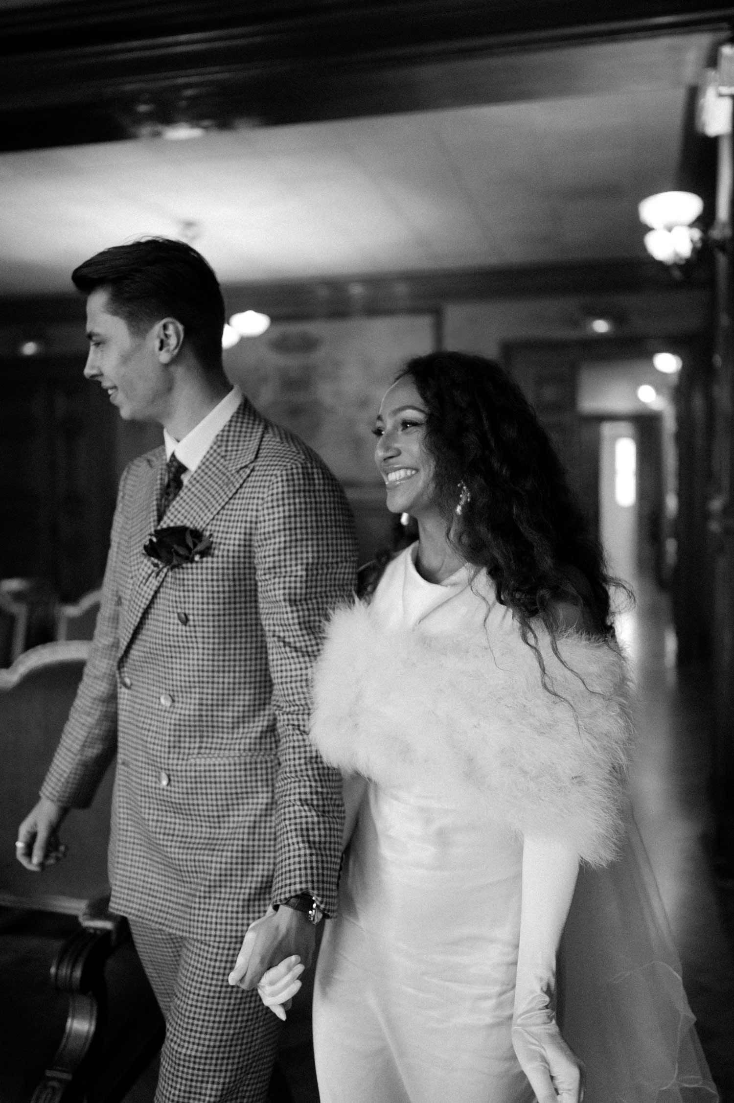 A black and white photo of a couple walking through an elegant vintage hallway.