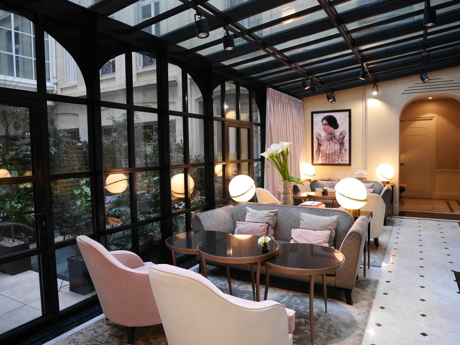 Bright Parisian hotel conservatory lounge with blush chairs, globe lights, and garden views.