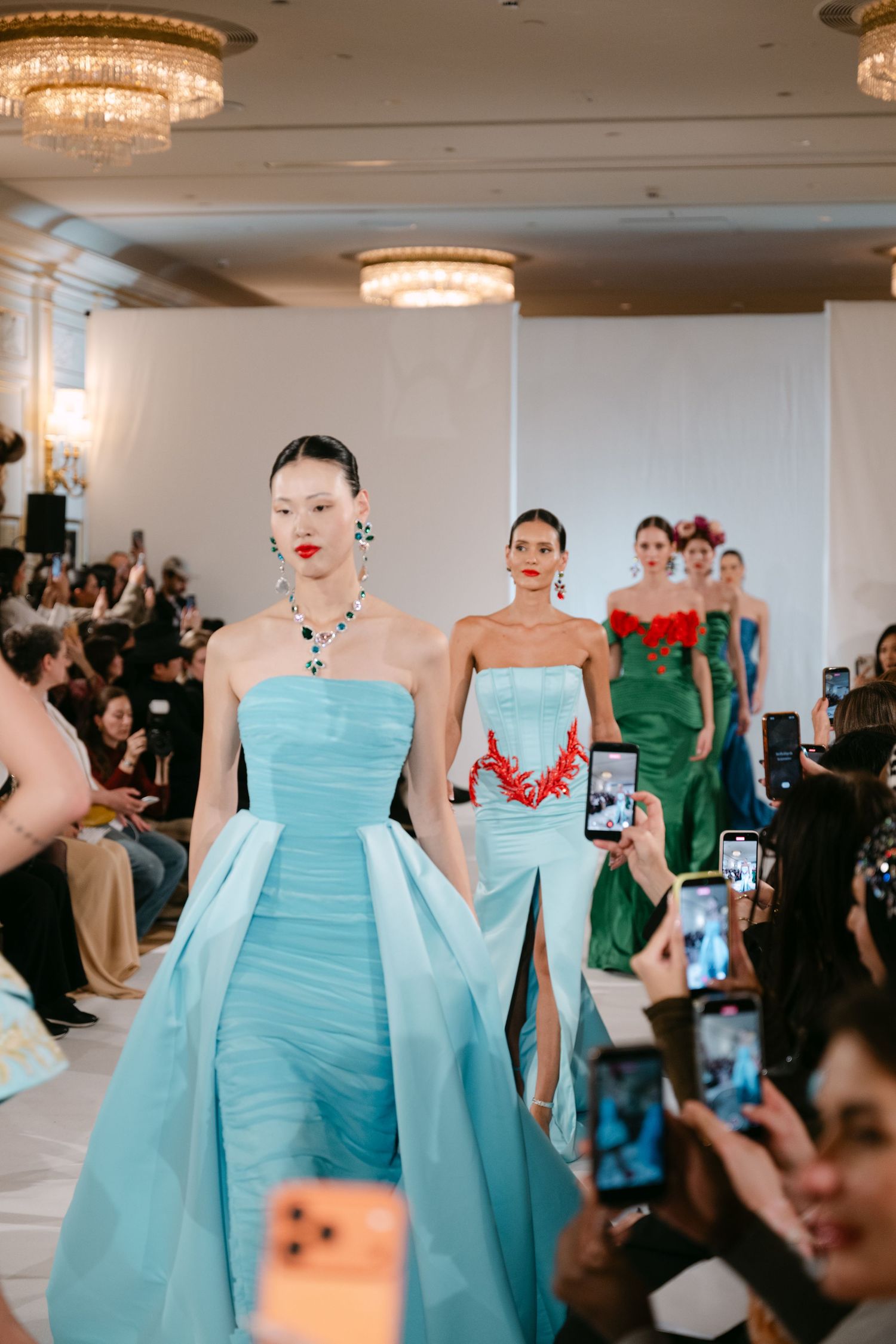 Models walk runway in blue and green strapless gowns at elegant couture fashion show.