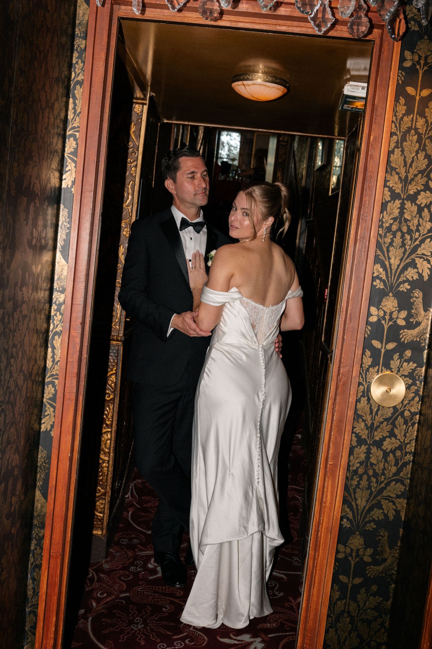 Bride and groom standing in a doorway with ornate gold wallpaper decor.