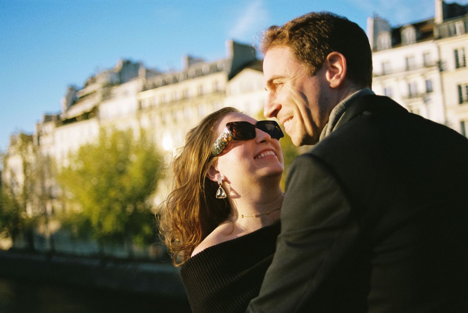 Couple sharing a romantic moment in Paris, woman wearing sunglasses, with classic Haussmann buildings in the background.