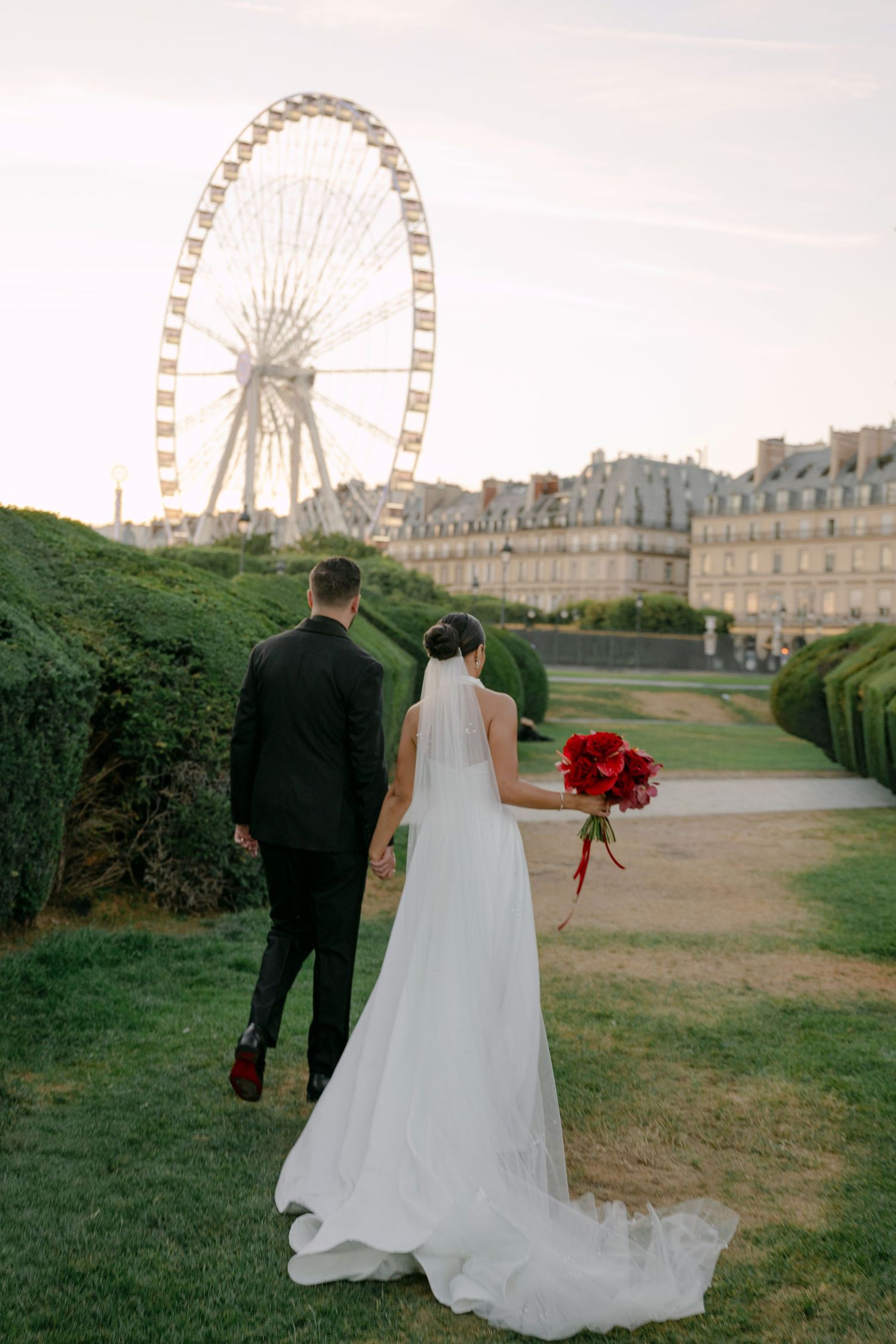 Bride and groom walk hand-in-hand in Tuileries Garden, Paris, with a Ferris wheel and red bouquet in view.