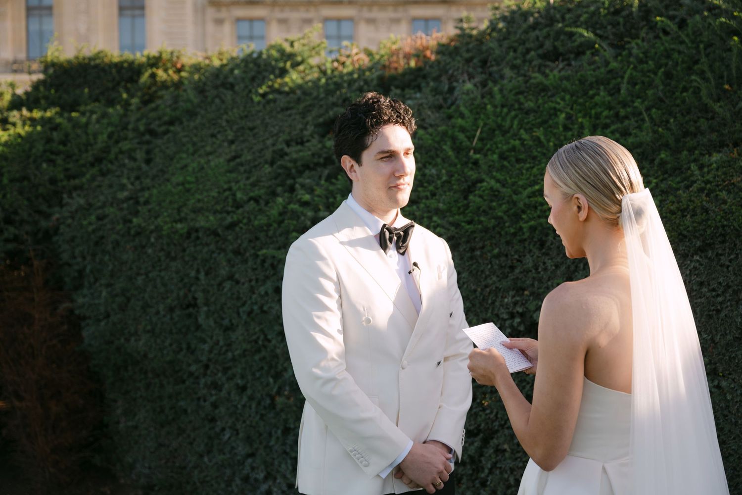 Couple exchanging vows outdoors at elegant garden wedding, groom in white suit, bride in sleeveless gown reading vows.