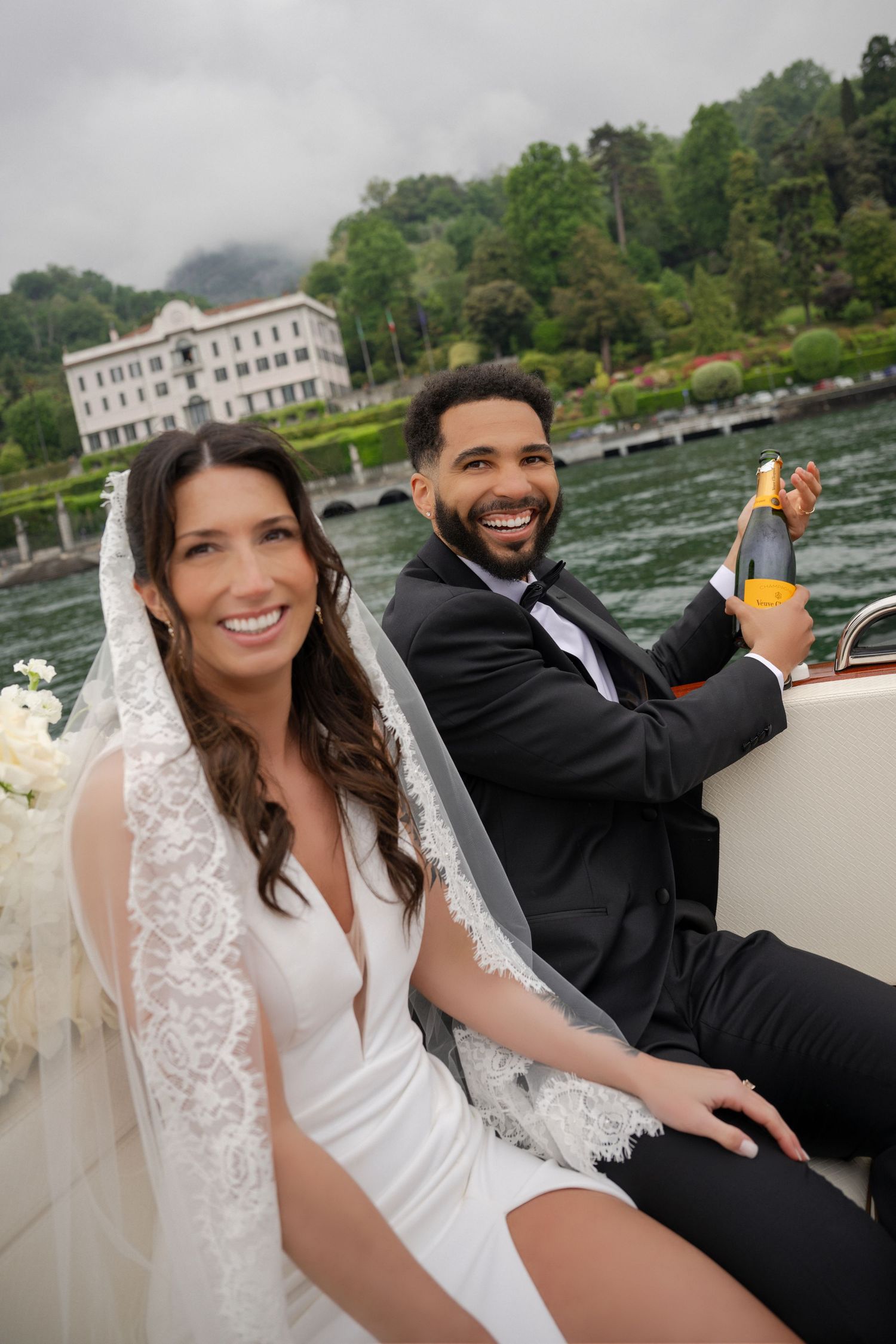 Smiling bride and groom celebrate on a boat with champagne, elegant villa visible on lakeside behind them.
