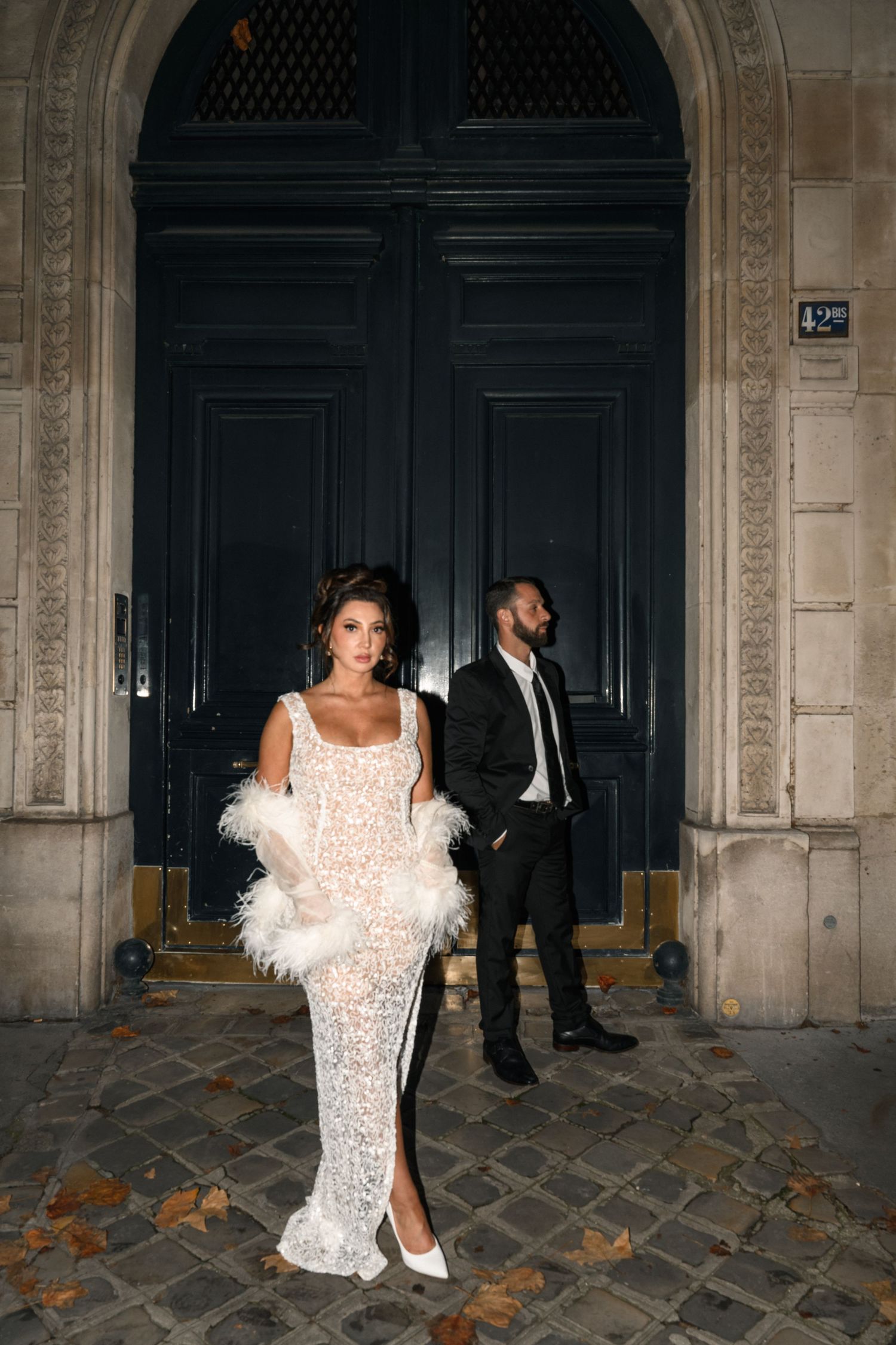 Elegant bride in lace gown with feather wrap stands before grand dark Parisian doorway with groom behind her.