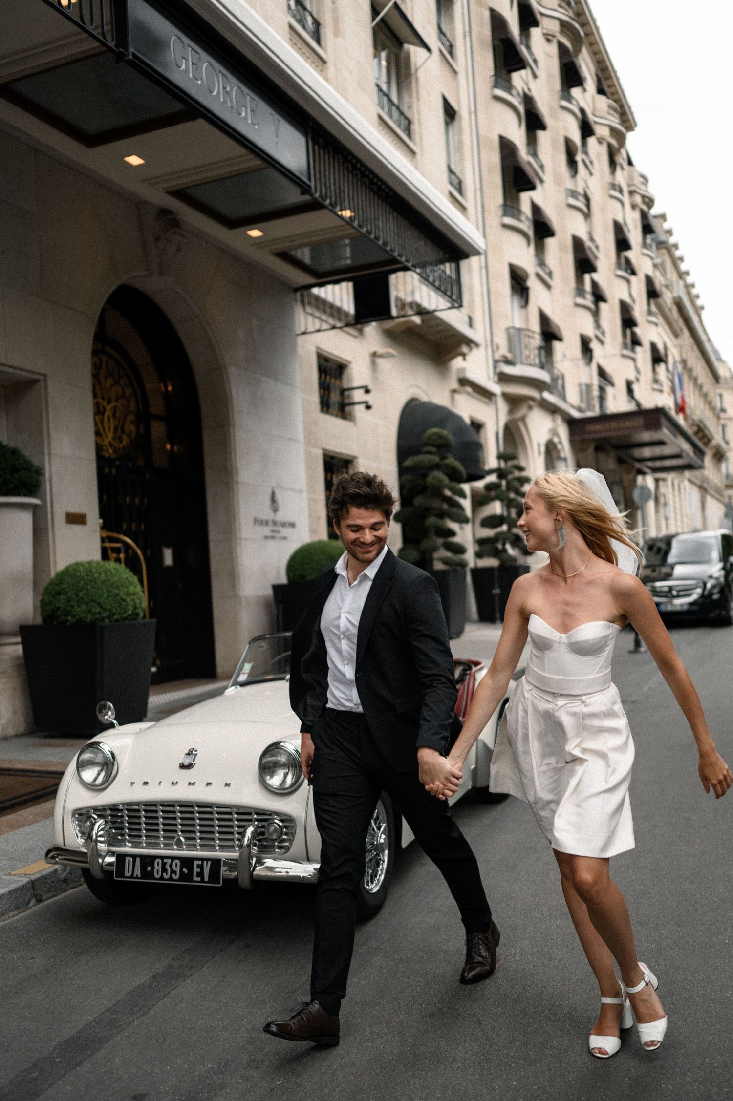 Bride and groom holding hands laughing outside a luxury Paris hotel with a vintage white classic car.