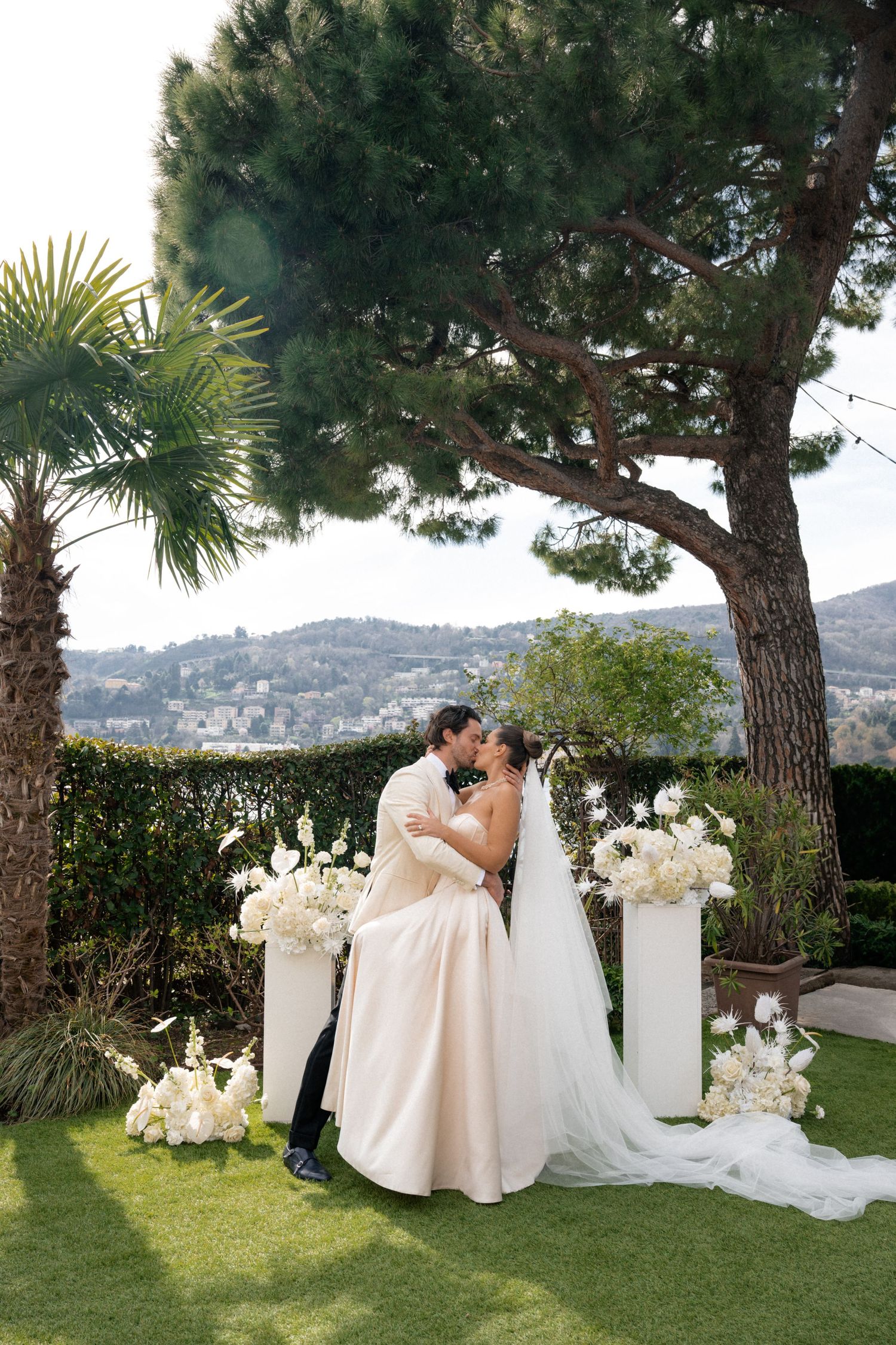Bride and groom share first kiss at outdoor wedding ceremony with white floral arrangements and scenic lake views.