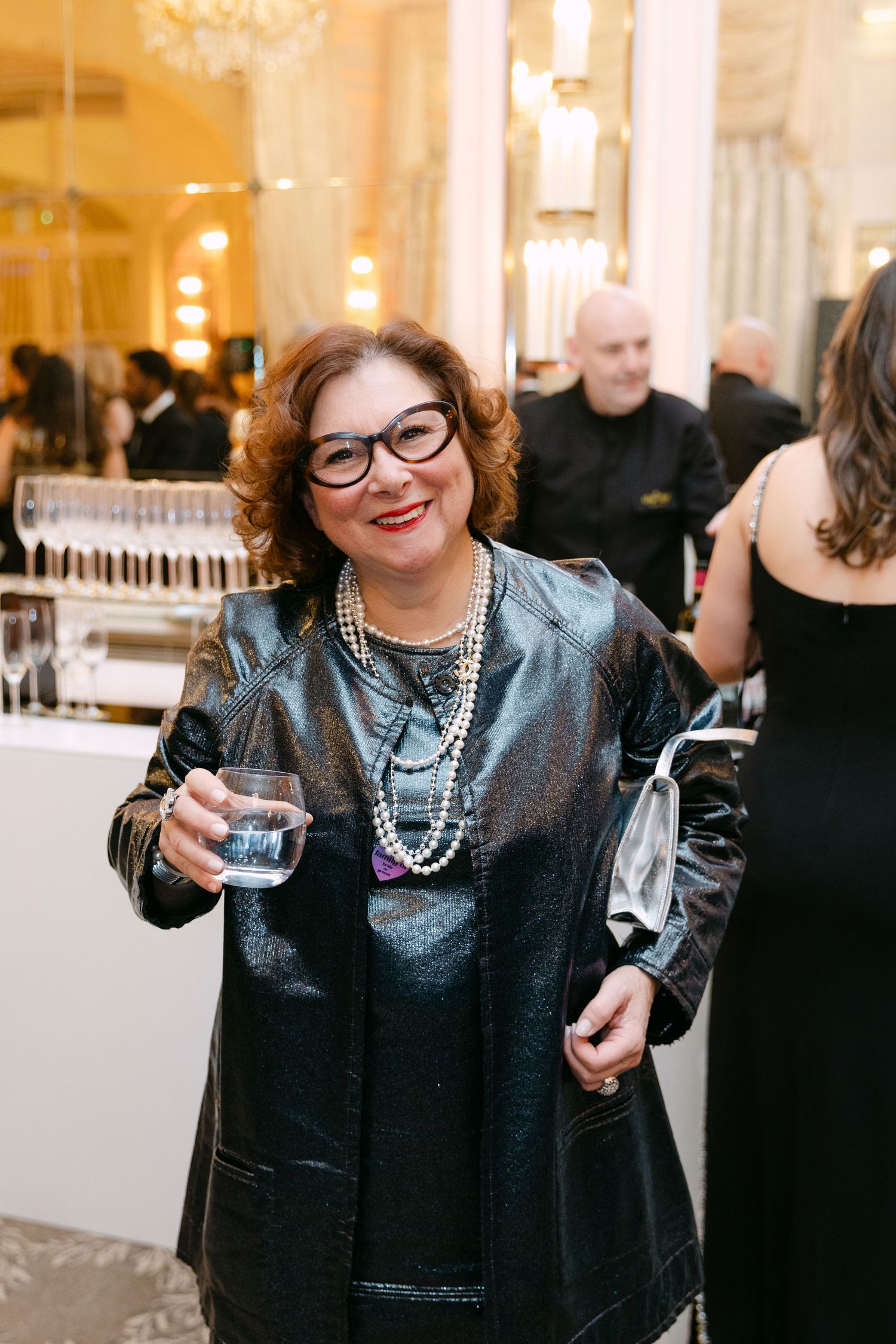 A smiling woman in a black leather jacket and pearl necklaces holds a water glass at an elegant indoor event.