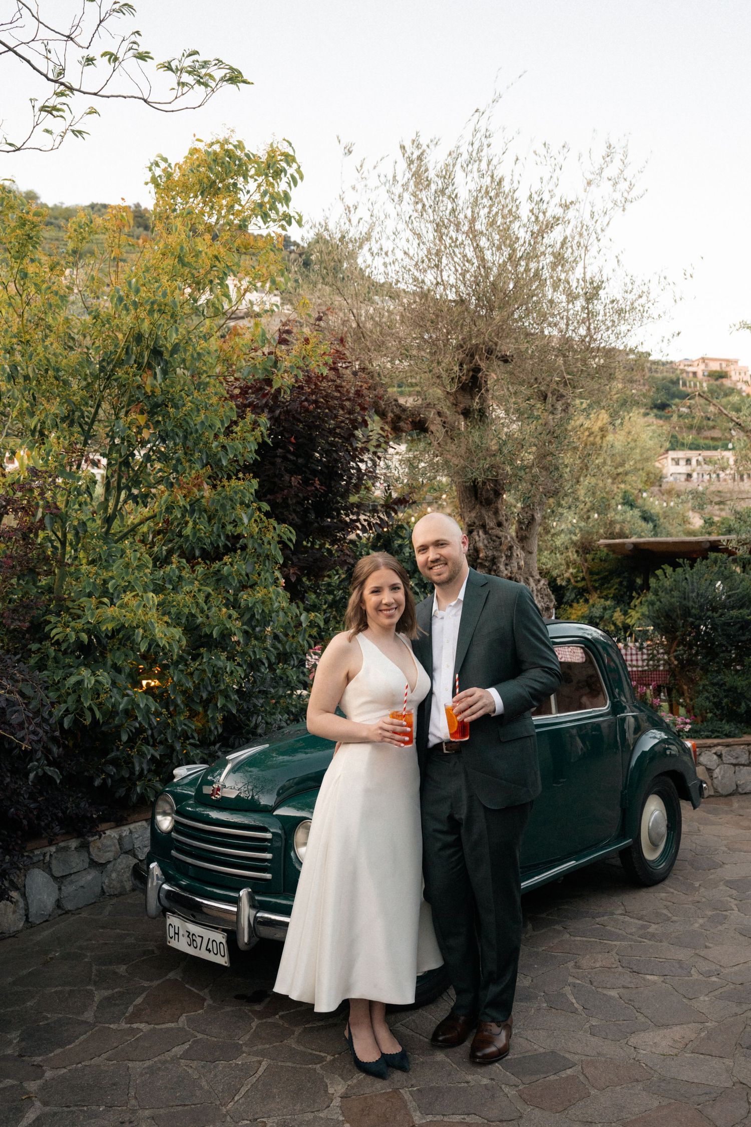 Smiling bride and groom pose with Aperol Spritz beside a vintage green Fiat in Italian garden setting.