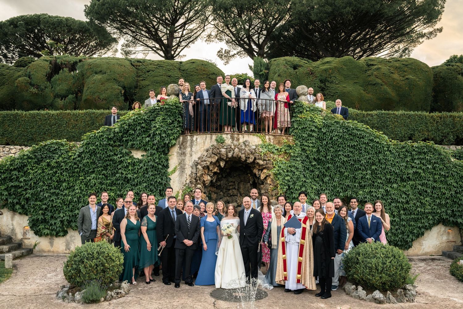 Large wedding party group photo on terraced garden steps surrounded by manicured hedges.