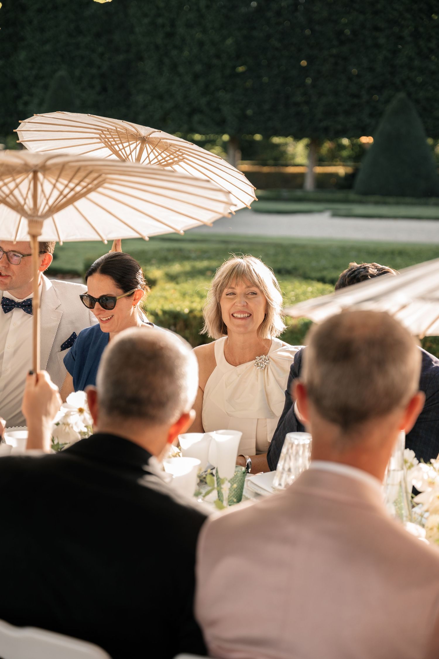 Wedding guests smiling and socializing at an outdoor reception under white parasol umbrellas.