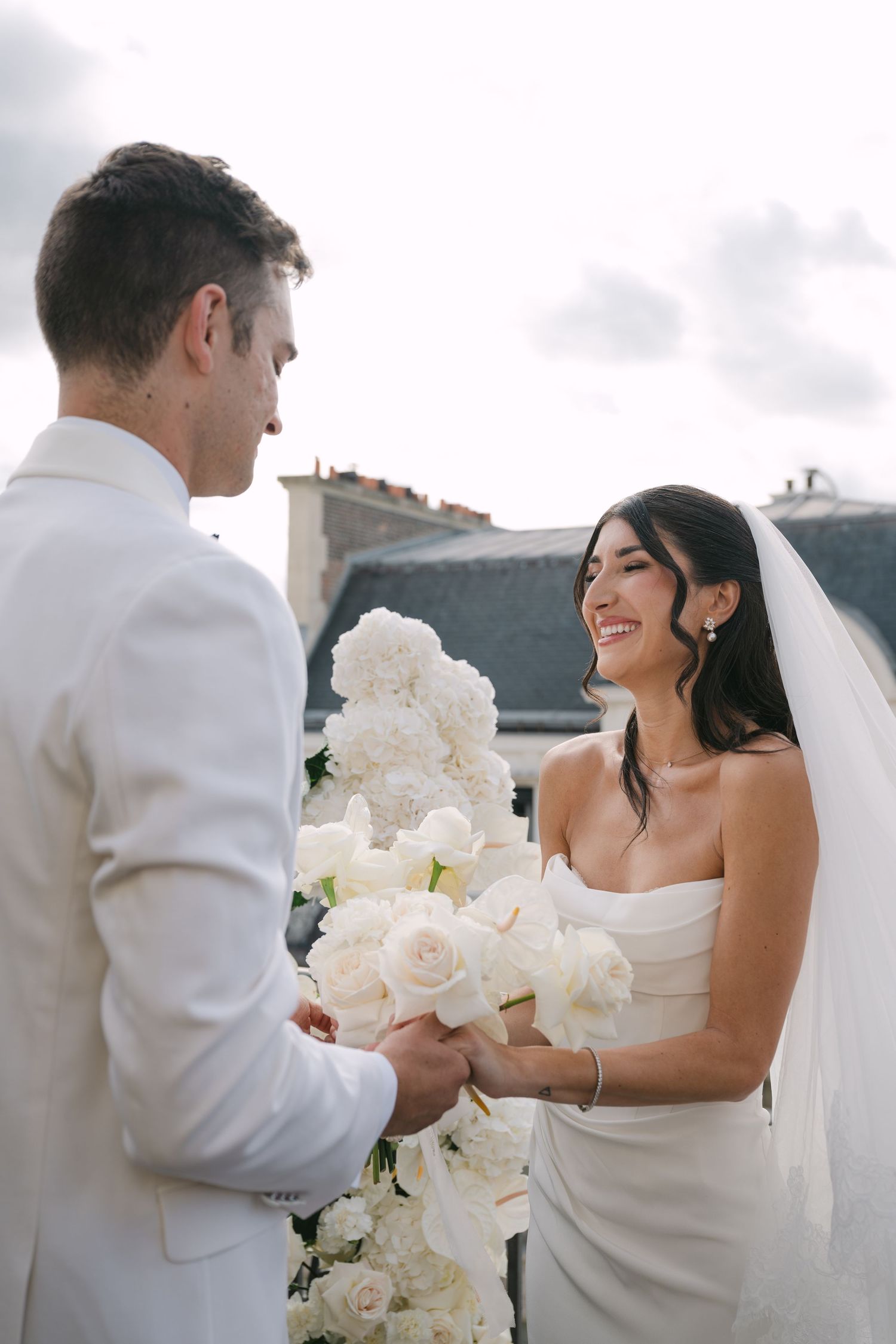 Smiling bride in strapless white gown holds hands with groom at outdoor rooftop wedding ceremony.