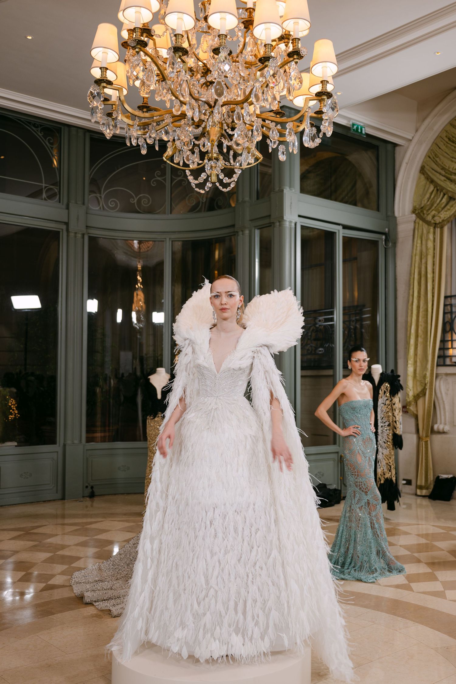 Model wears white feathered gown with dramatic ruffled collar in elegant chandelier ballroom.